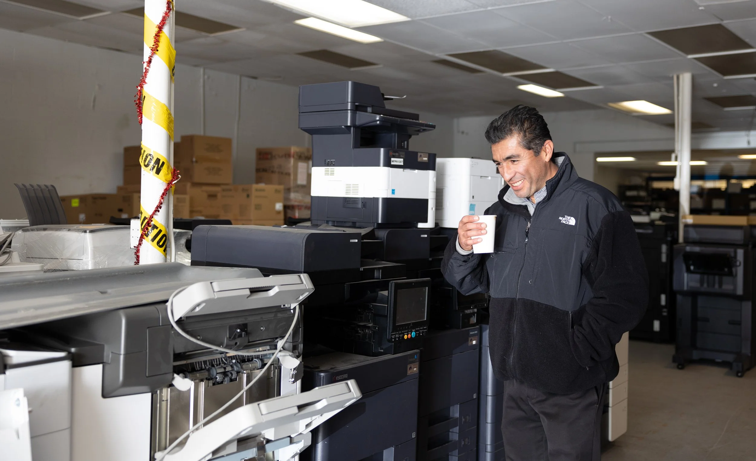 Man smiling holding a paper cup standing in front of office printers and copiers in a warehouse or office supply room.