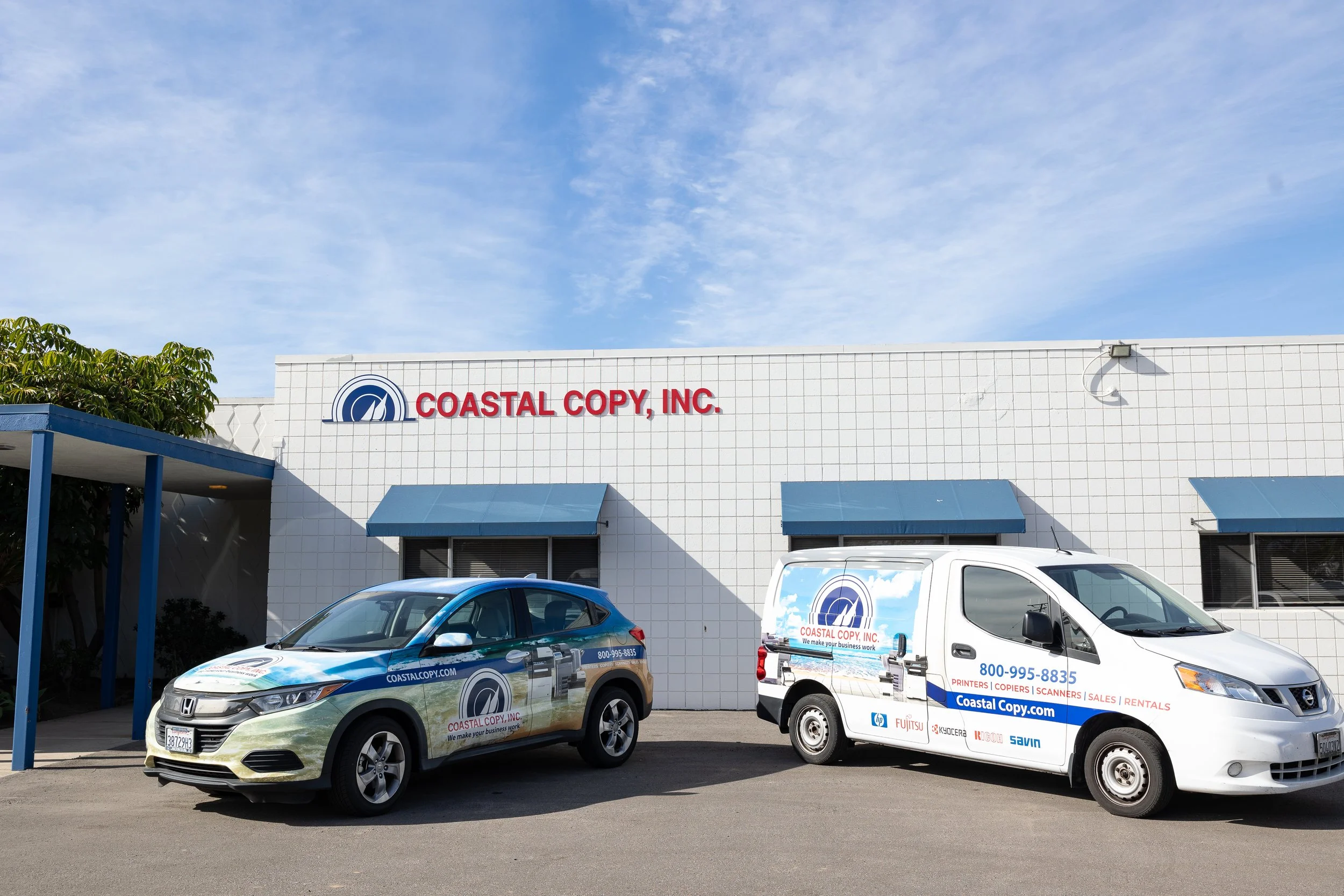Exterior of Coastal Copy Inc. building with two vehicles parked in front, one van and one SUV, both branded with the company's logo and contact information.