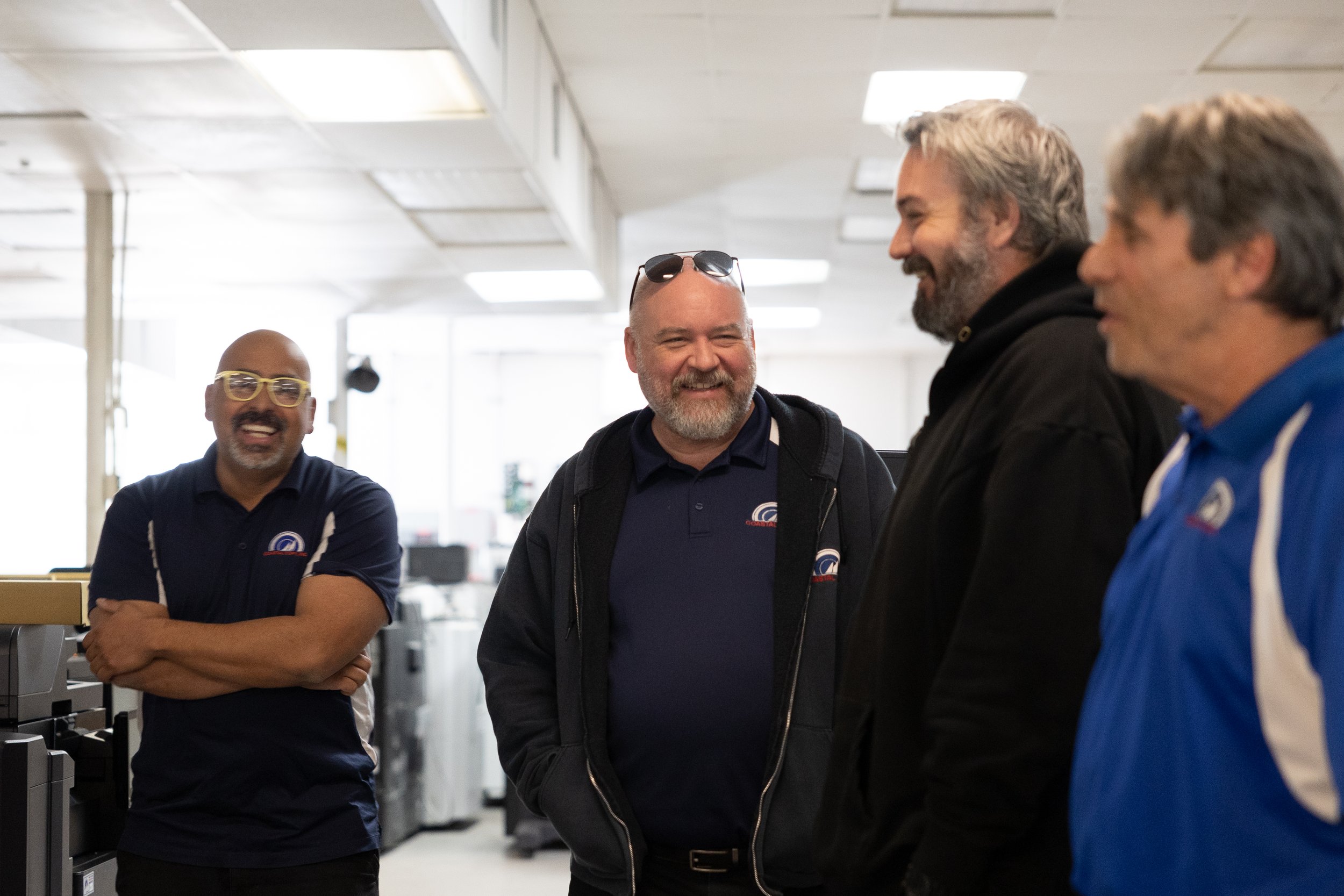 Four men are smiling and conversing inside an office or store with bright overhead lighting. One man is wearing yellow glasses and a navy blue polo shirt, another man is wearing sunglasses on his head and a navy blue jacket, the third man is in a black hoodie, and the fourth man is in a blue jacket with white accents.