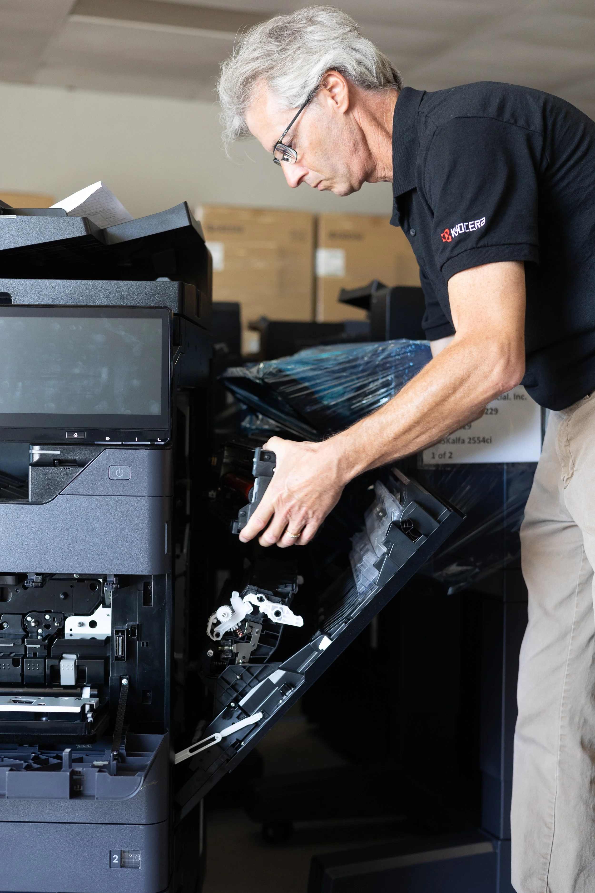 A man with gray hair and glasses repairing a photocopier or printer in an office or warehouse setting.