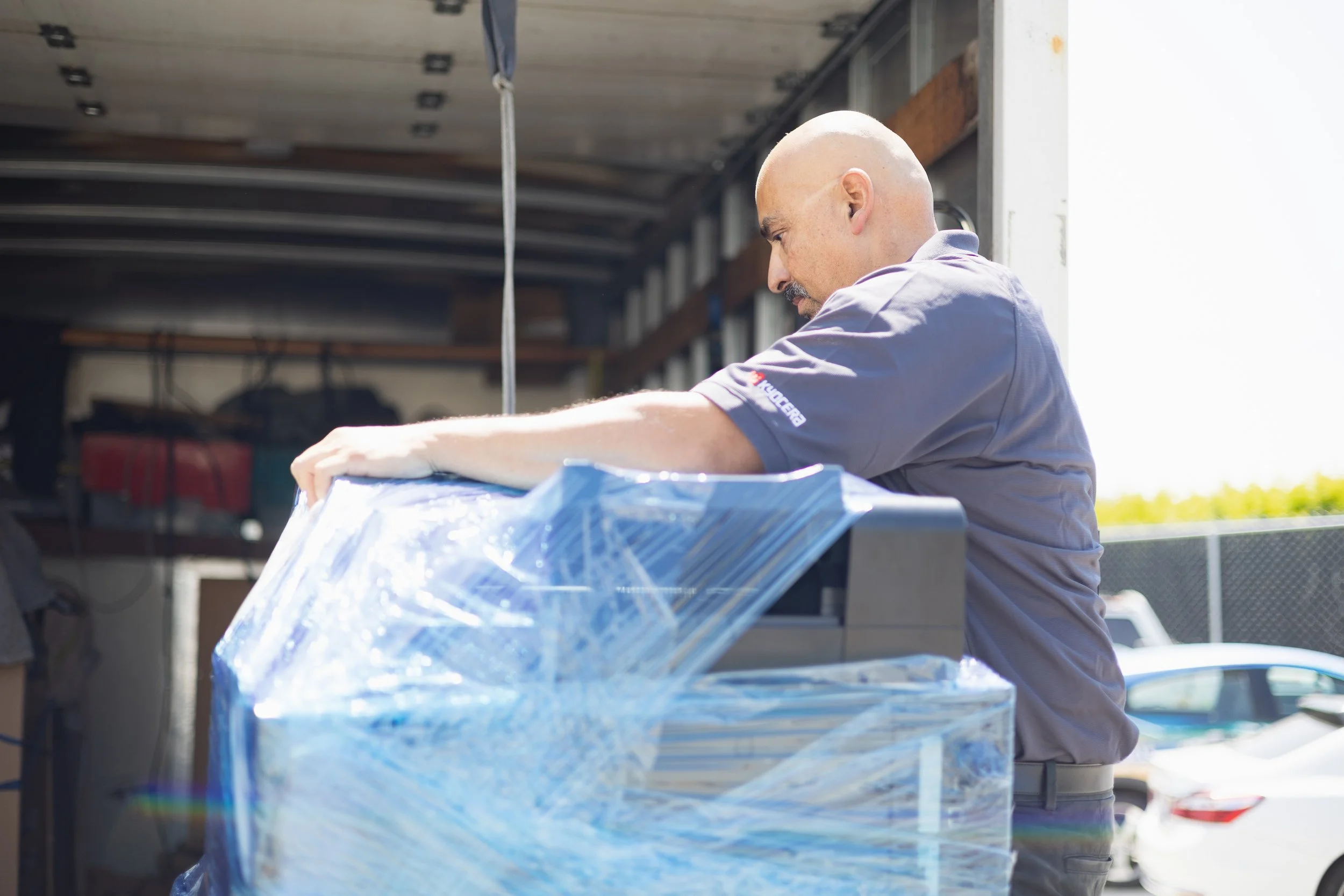 A man with a bald head is handling a large wrapped item outside near a delivery truck.