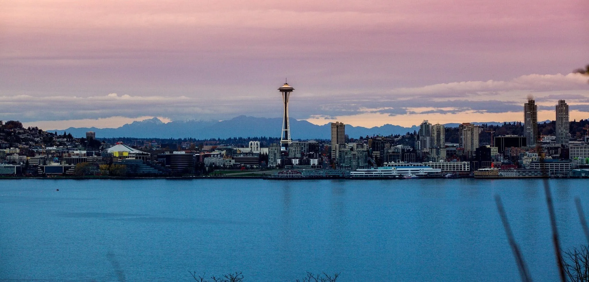 View of Seattle skyline at dusk with the Space Needle in the center, mountains in the background, and a body of water in the foreground.