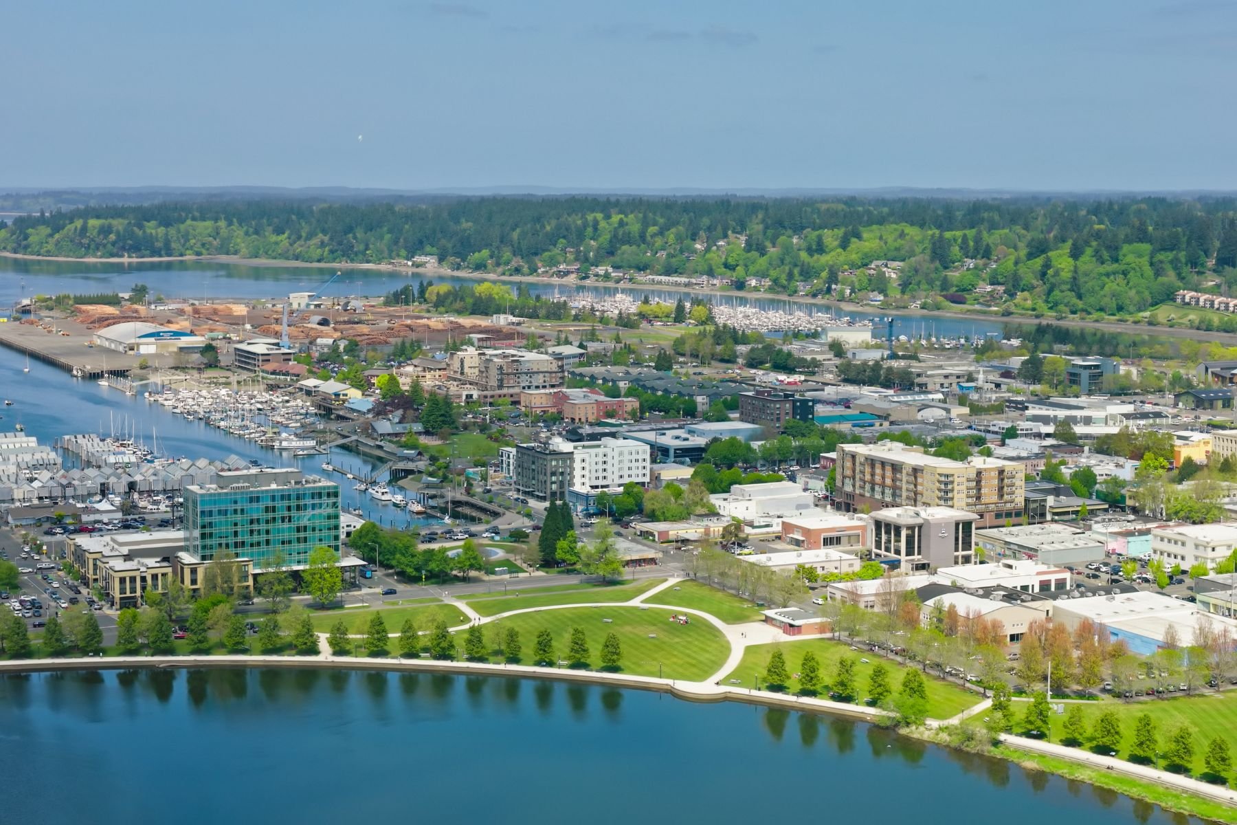 Aerial view of a city with a river and marina, green parks, and a mixture of commercial and residential buildings in a lush, hilly landscape.