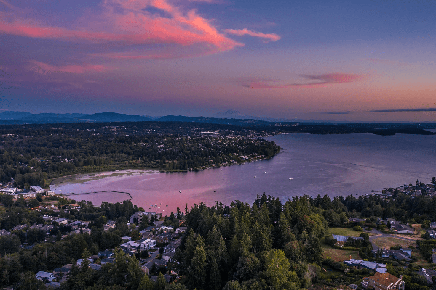 Aerial view of a town along a river during sunset, with pink and purple sky and mountains in the background.