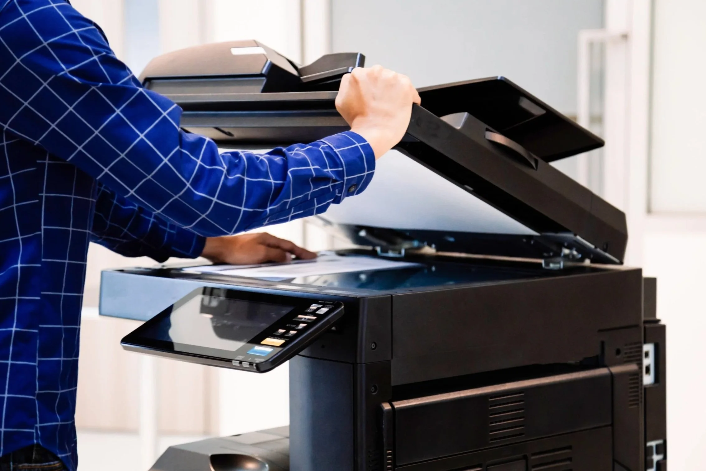 Person in a blue checkered shirt using a large multifunction printer or copier.