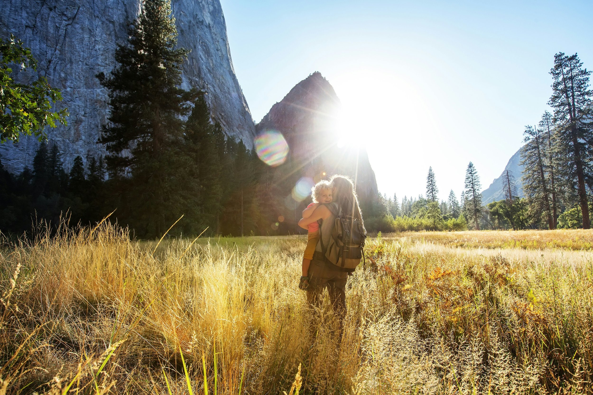 A person carrying a child in a natural landscape with tall golden grass, tall pine trees, and large rocky mountains under a clear bright sky, with the sun shining directly.
