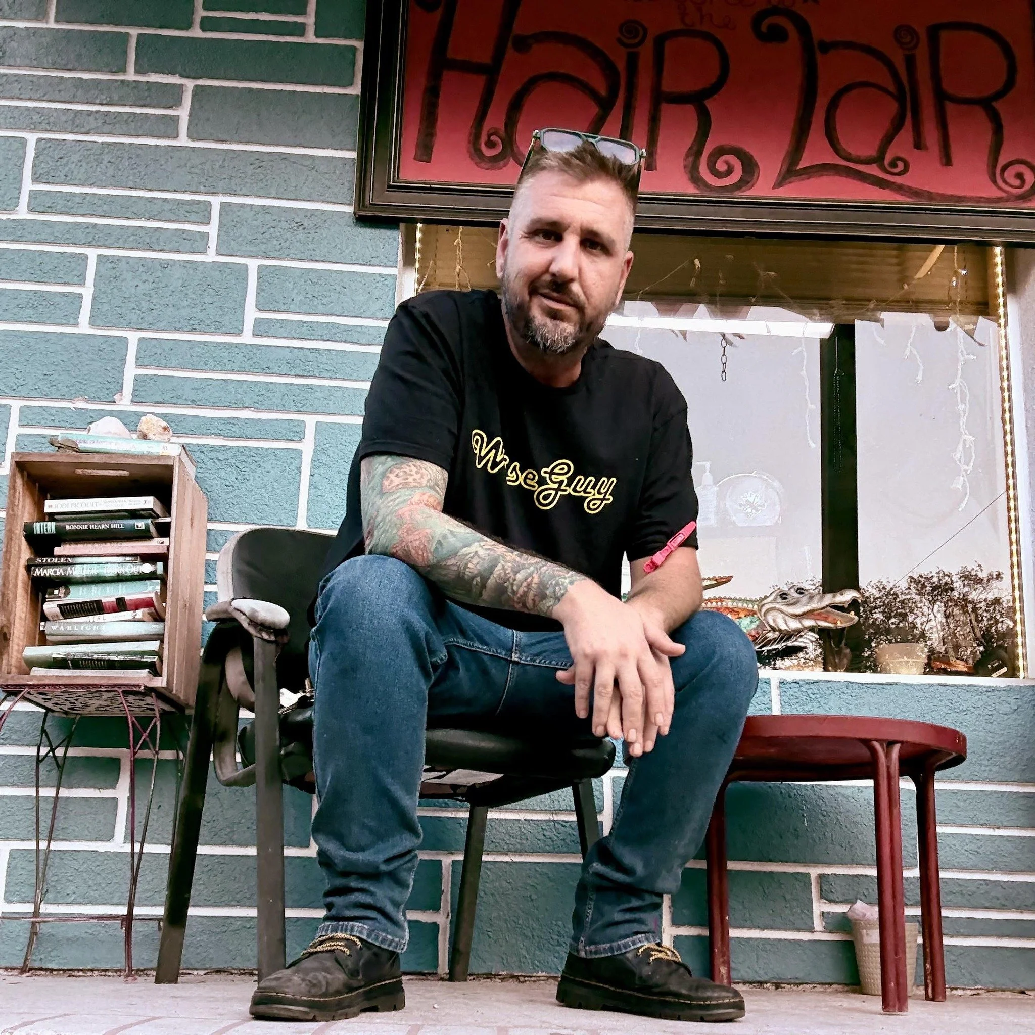 A man sitting in front of a brick wall outside a storefront that reads "Hair Lab." He has short hair, a beard, tattoos on his arms, and is wearing glasses on his head. He's dressed in a black T-shirt with yellow text, jeans, and black boots.