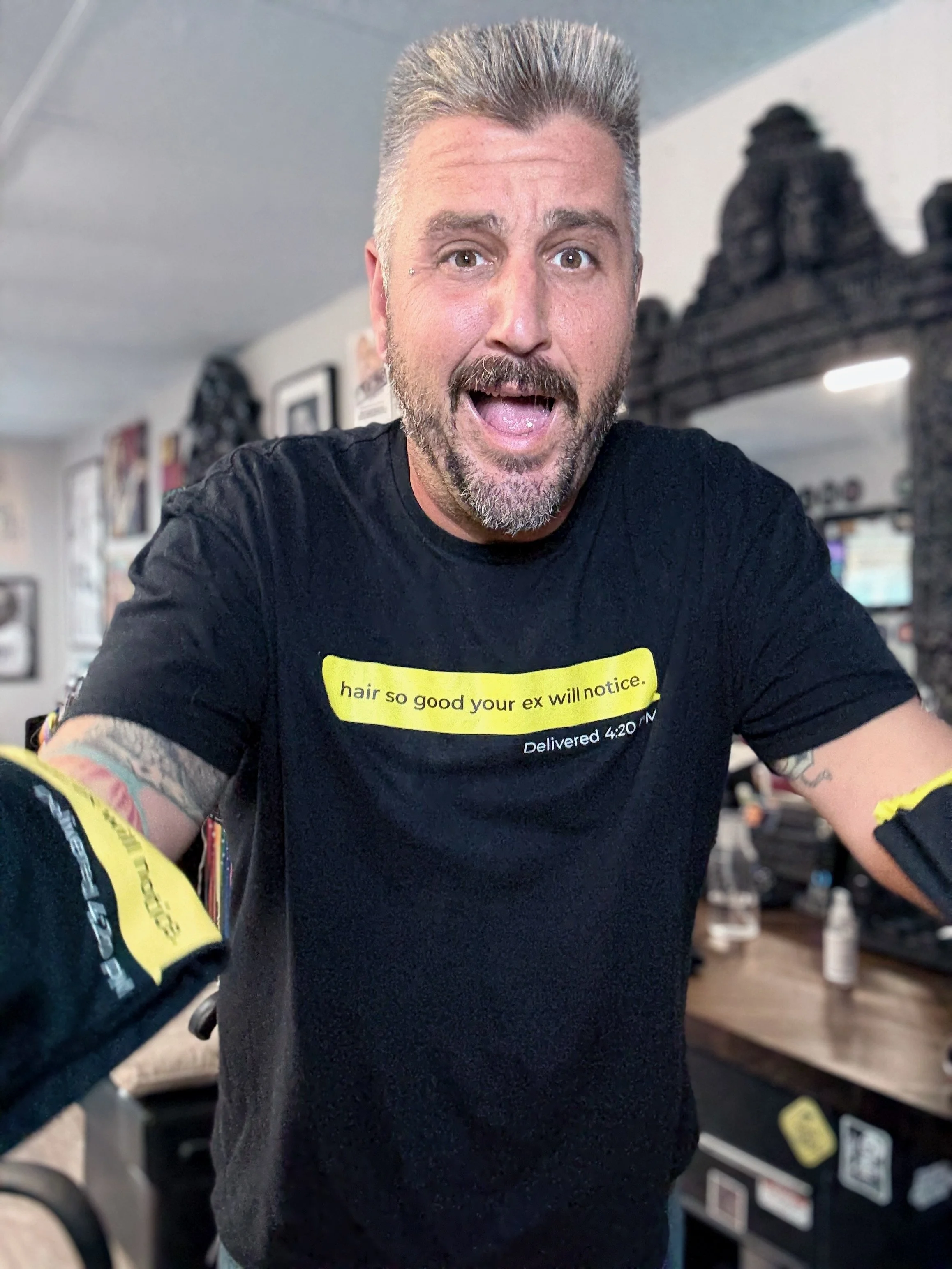 A man with gray hair and a beard, wearing a black T-shirt with yellow text, is smiling and reaching towards the camera. The background shows a room with various framed pictures, a mirror, and a wooden table with bottles and supplies.