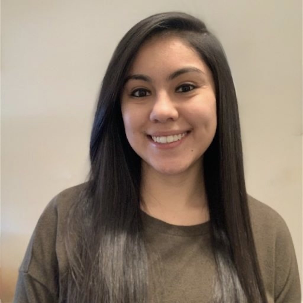 Portrait of a young woman with long dark hair, smiling, wearing a casual brown top, standing against a plain light-colored background.