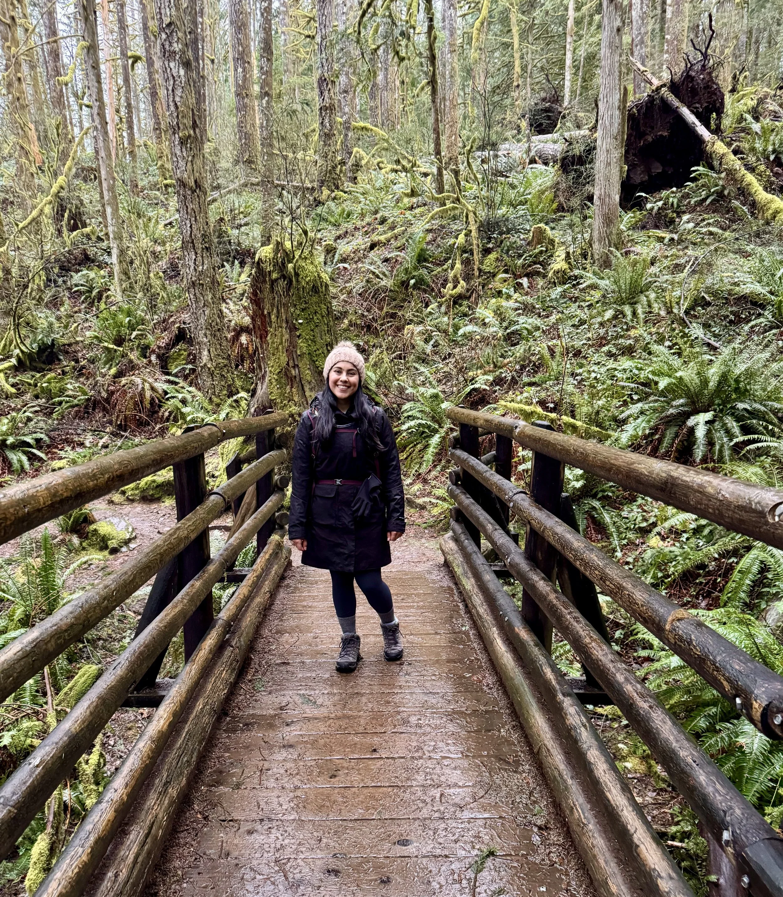 A woman standing on a wooden bridge surrounded by lush, green forest with moss-covered trees and ferns.