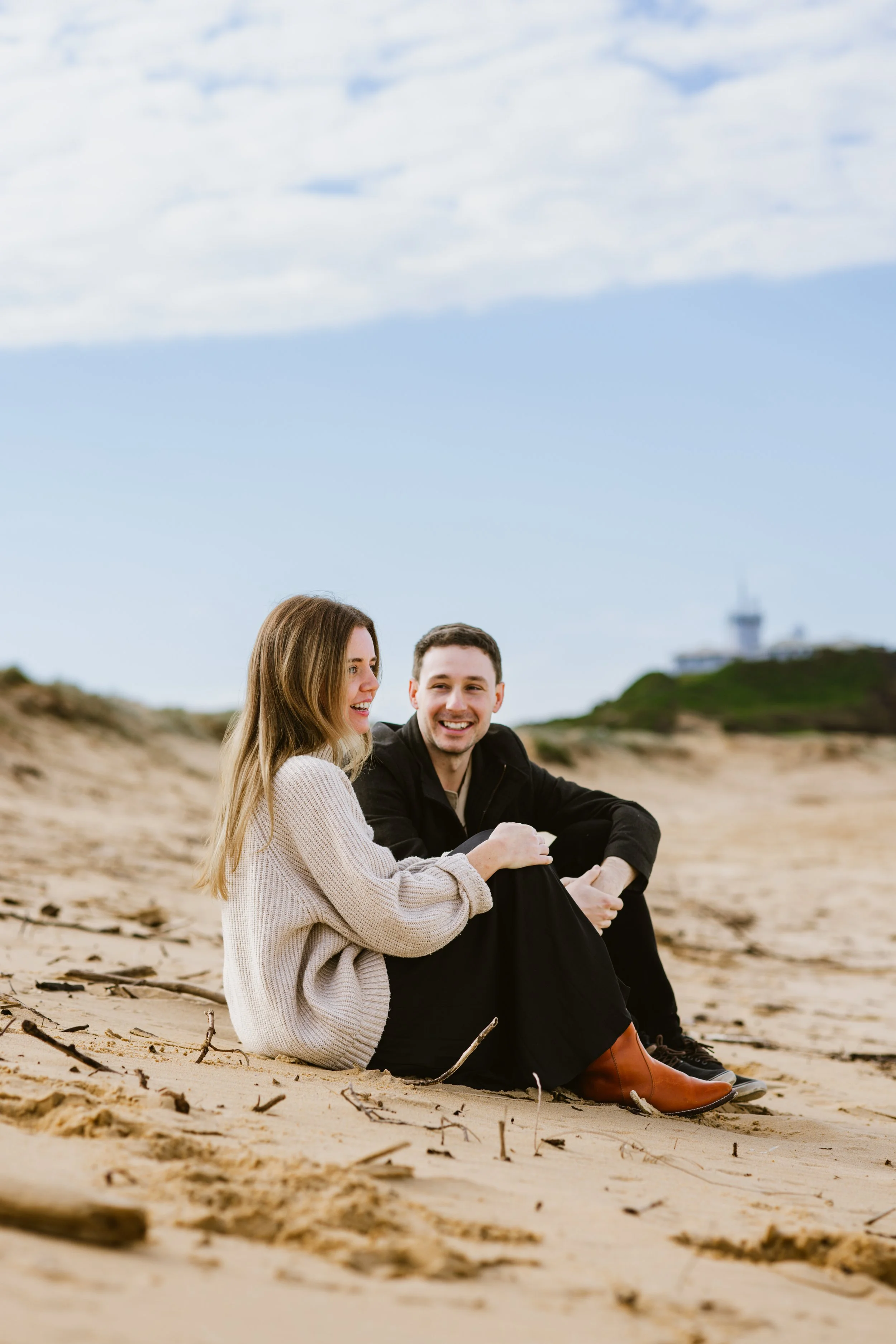 A smiling woman and man sitting on the sand at the beach, with a blue sky and clouds overhead and a distant building in the background.