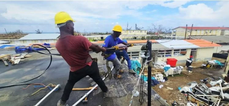 Two workers wearing yellow helmets working on a rooftop, one pulling a cable and the other operating a tool, with buildings and construction materials in the background.
