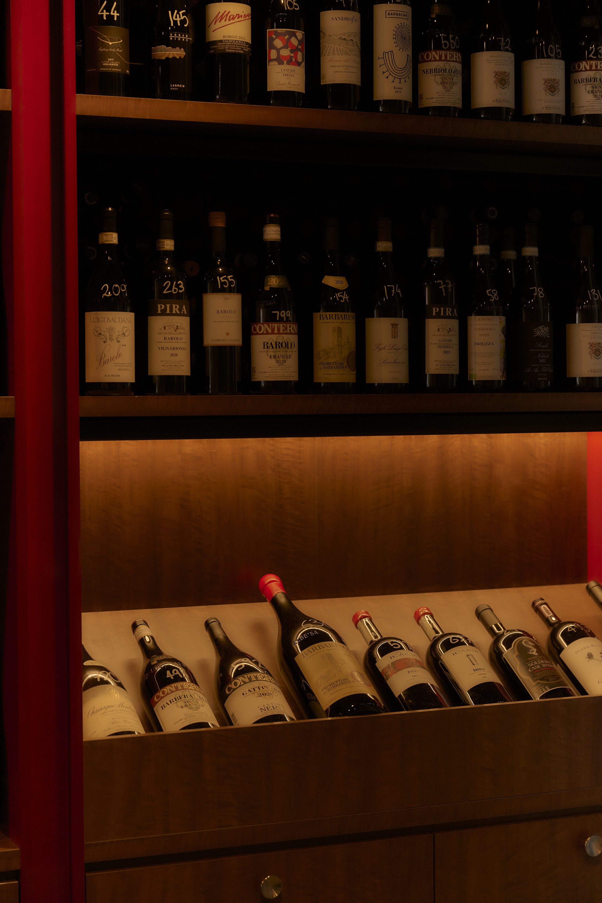 Wine bottles displayed on wooden shelves with labels visible, in a dimly lit environment.