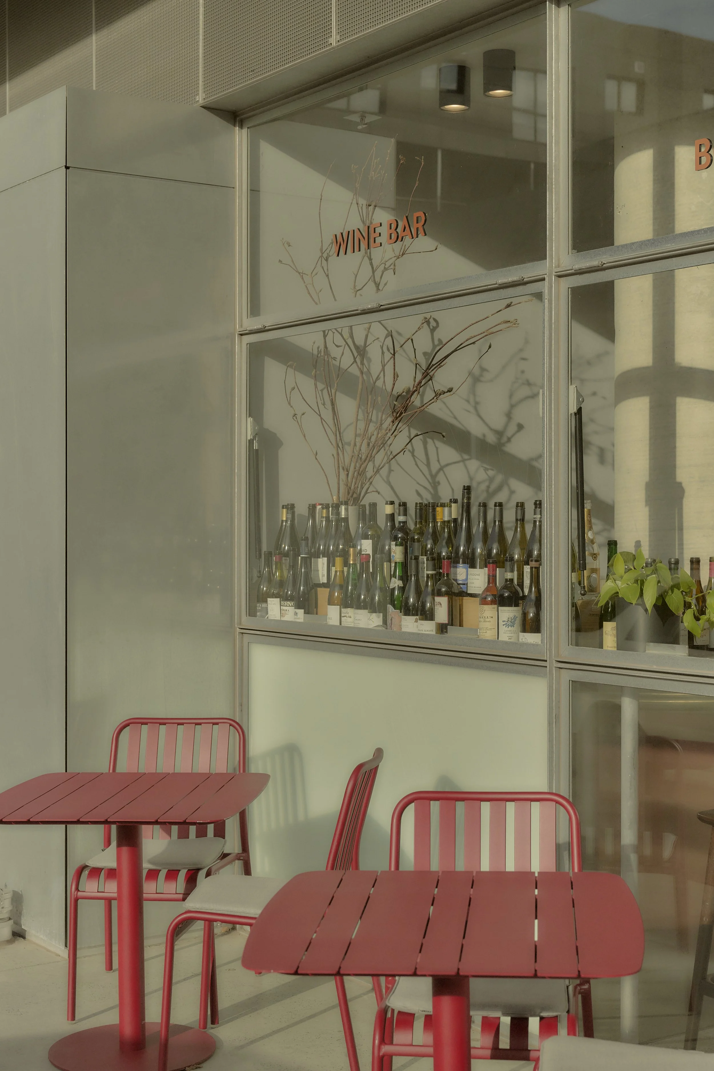 A windowed section of a wine bar with an assortment of wine bottles on display, a dry branch arrangement inside, red tables, and chairs outside.