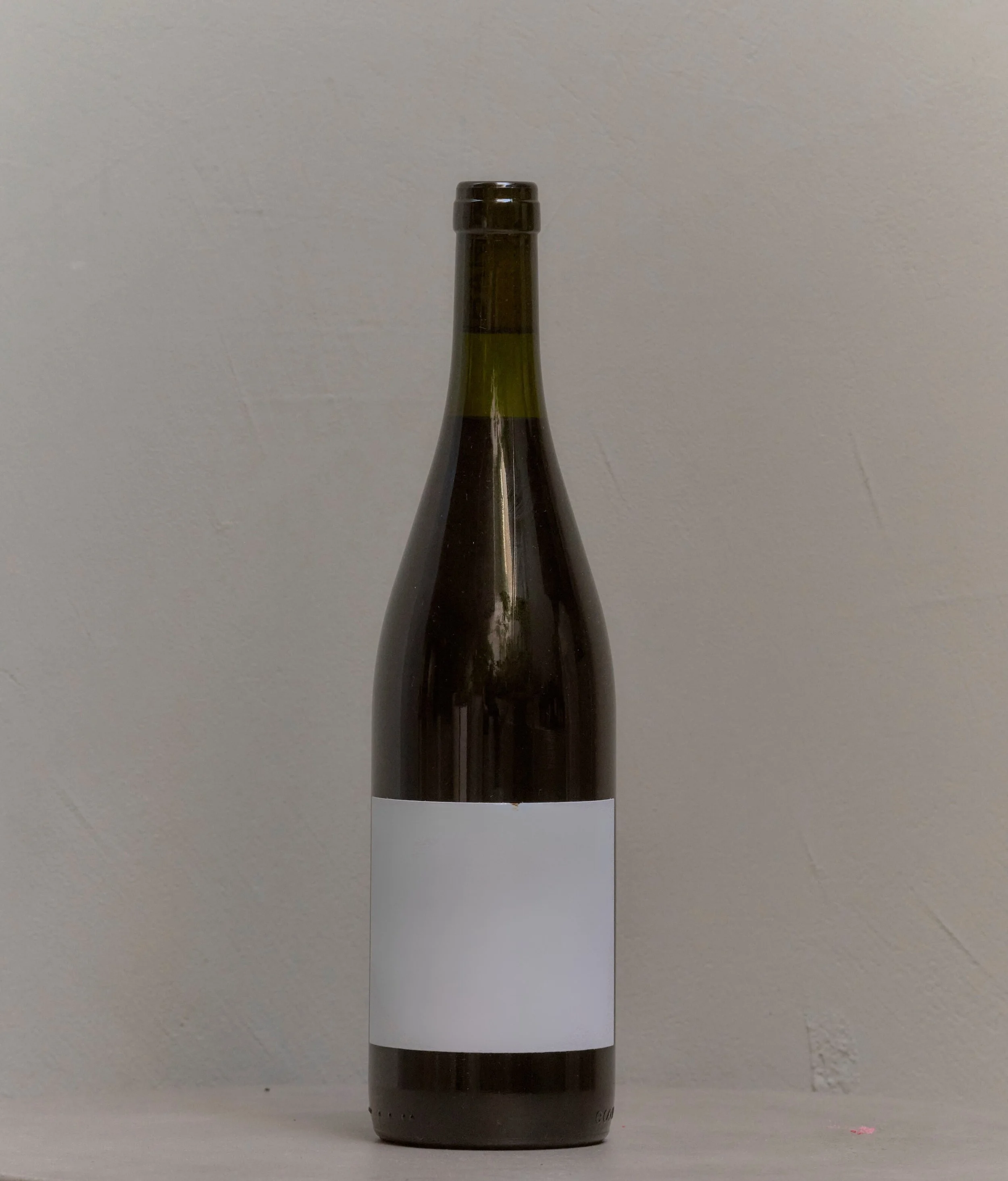 A dark glass wine bottle with a white blank label, standing on a white surface against a plain light-colored wall.