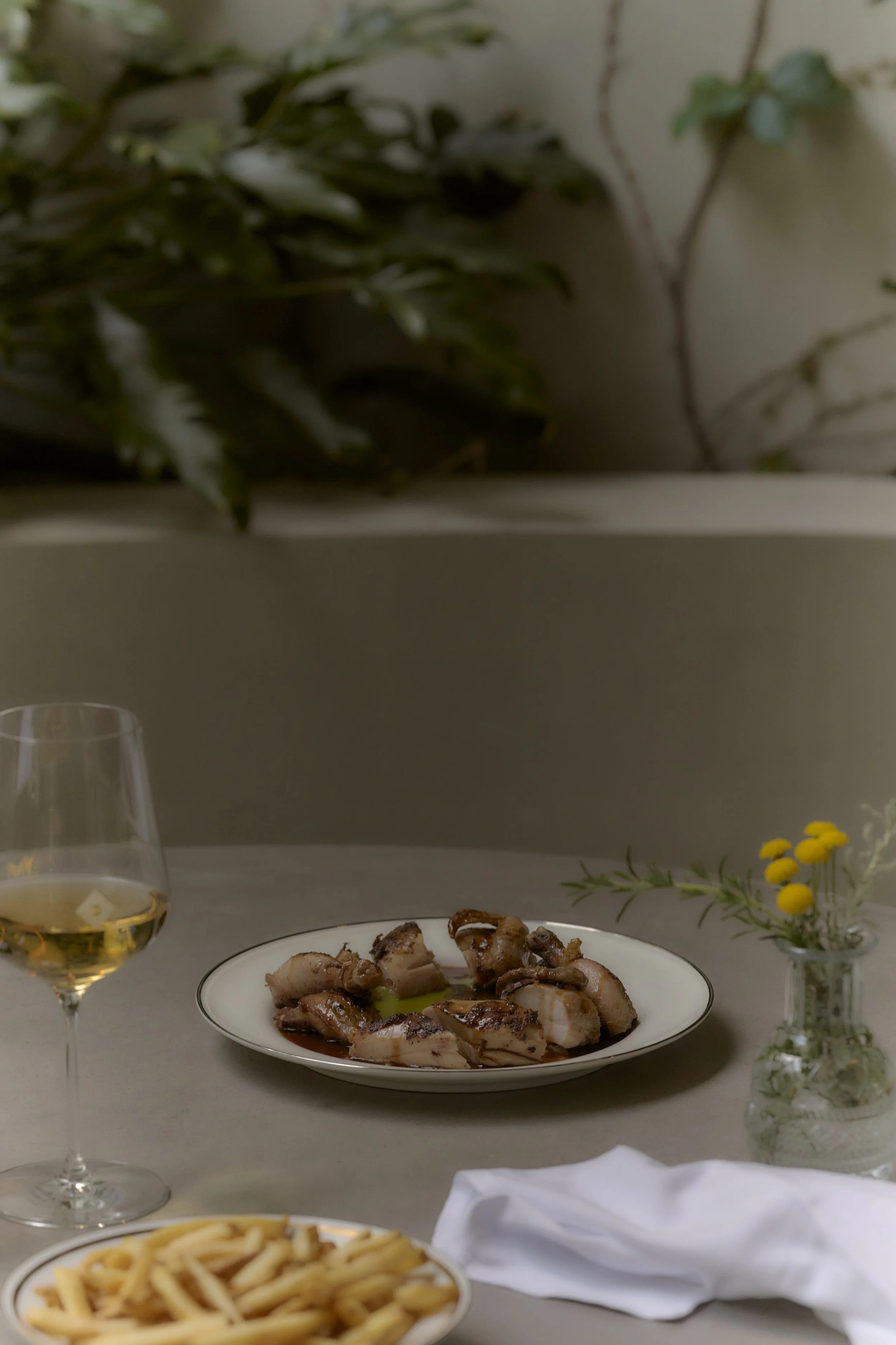 A plate of cooked octopus on a table with a glass of white wine, a bowl of spaghetti, a white cloth, and a small vase with yellow and white flowers, set against a background with green plants.