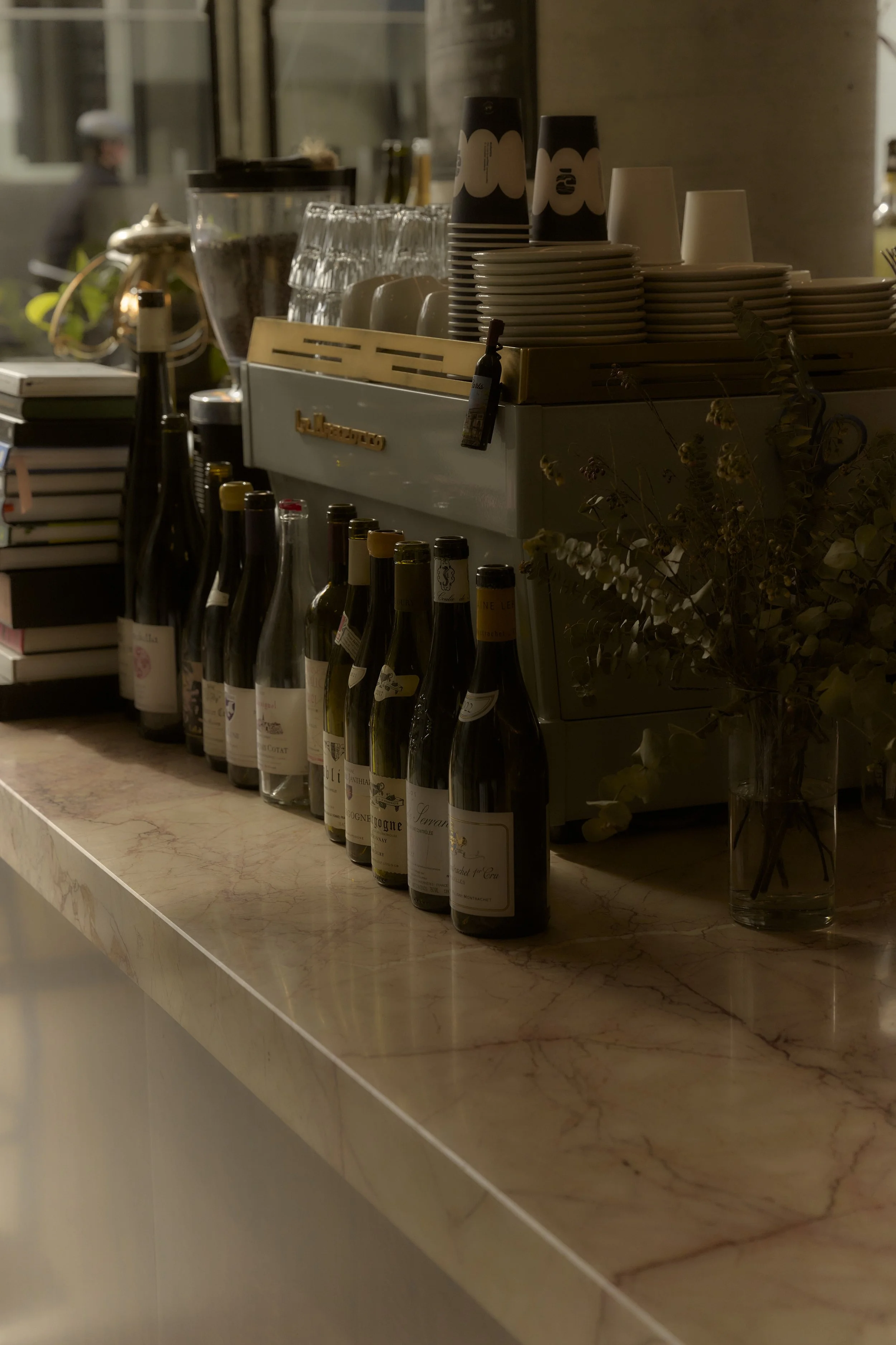 A row of wine bottles on a marble countertop next to a vase with dried flowers, with a coffee machine and stacks of cups and glasses in the background.