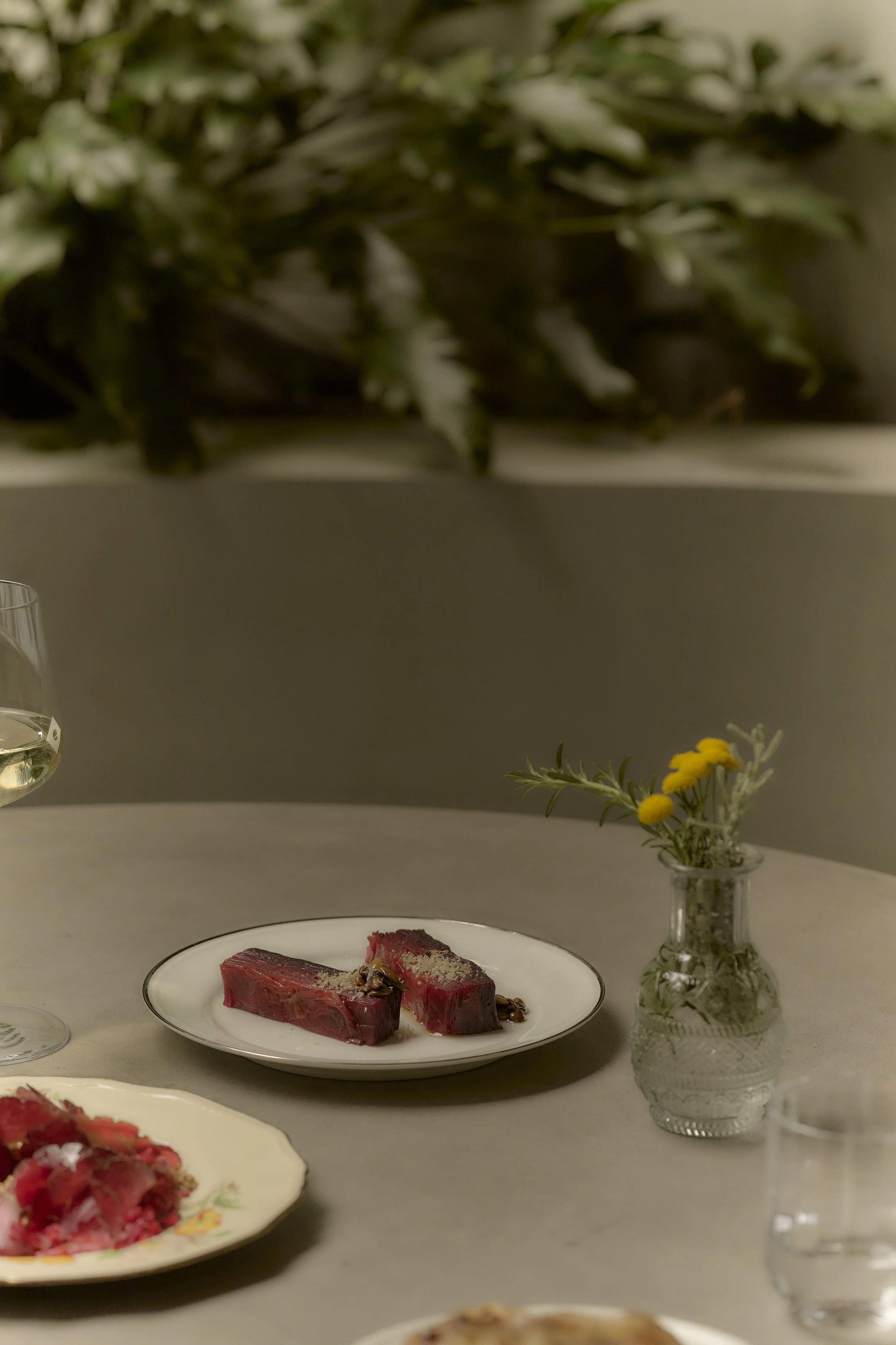 Two pieces of raw steak seasoned with salt on a white plate, decorated with herbs, on a kitchen or dining table with a vase of yellow flowers and glasses of water and wine in the background.