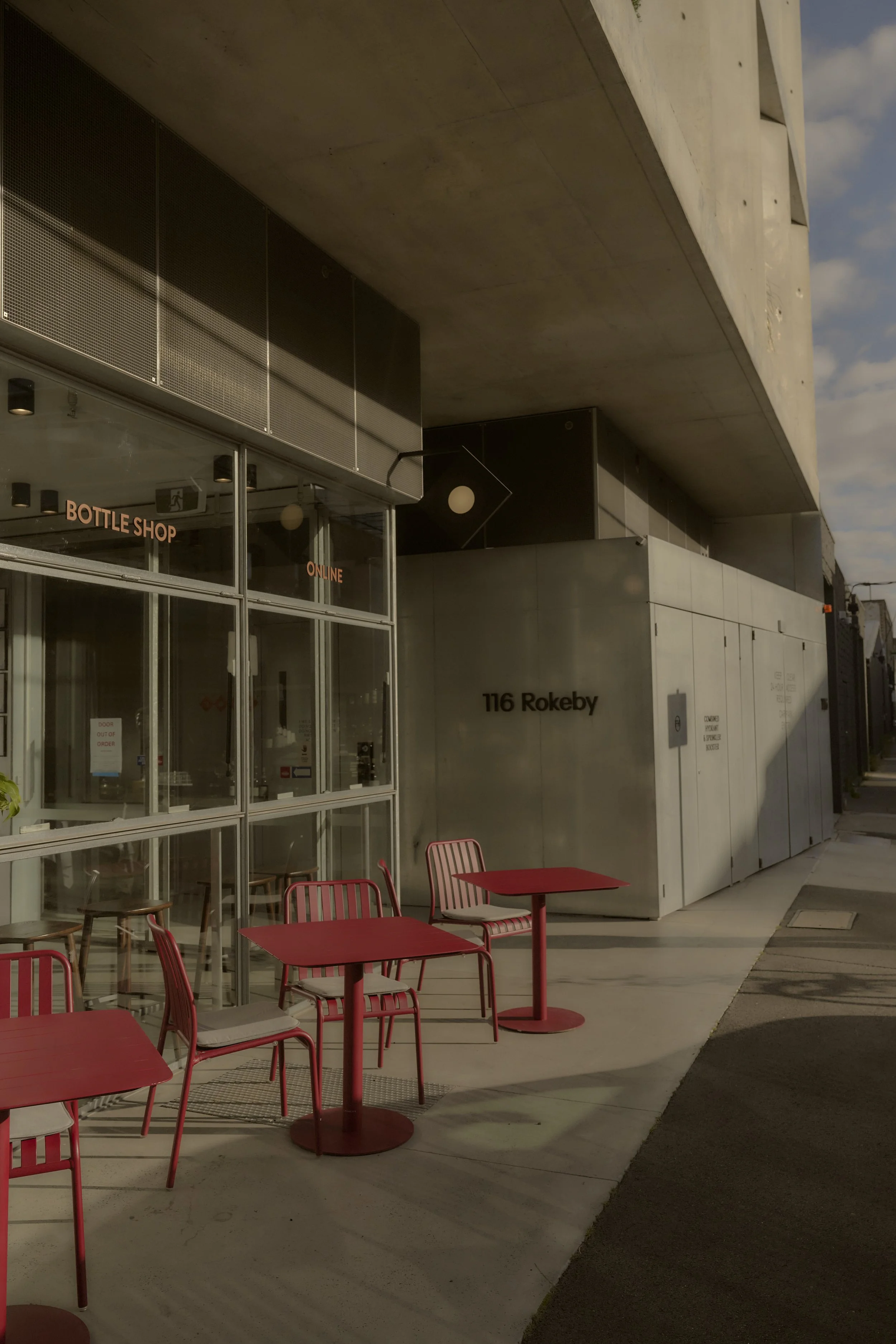 Outdoor seating area with red tables and chairs outside a modern building with glass windows and a silver wall. The building displays the address '116 Rokeby' and a sign indicating a bottle shop and online services.