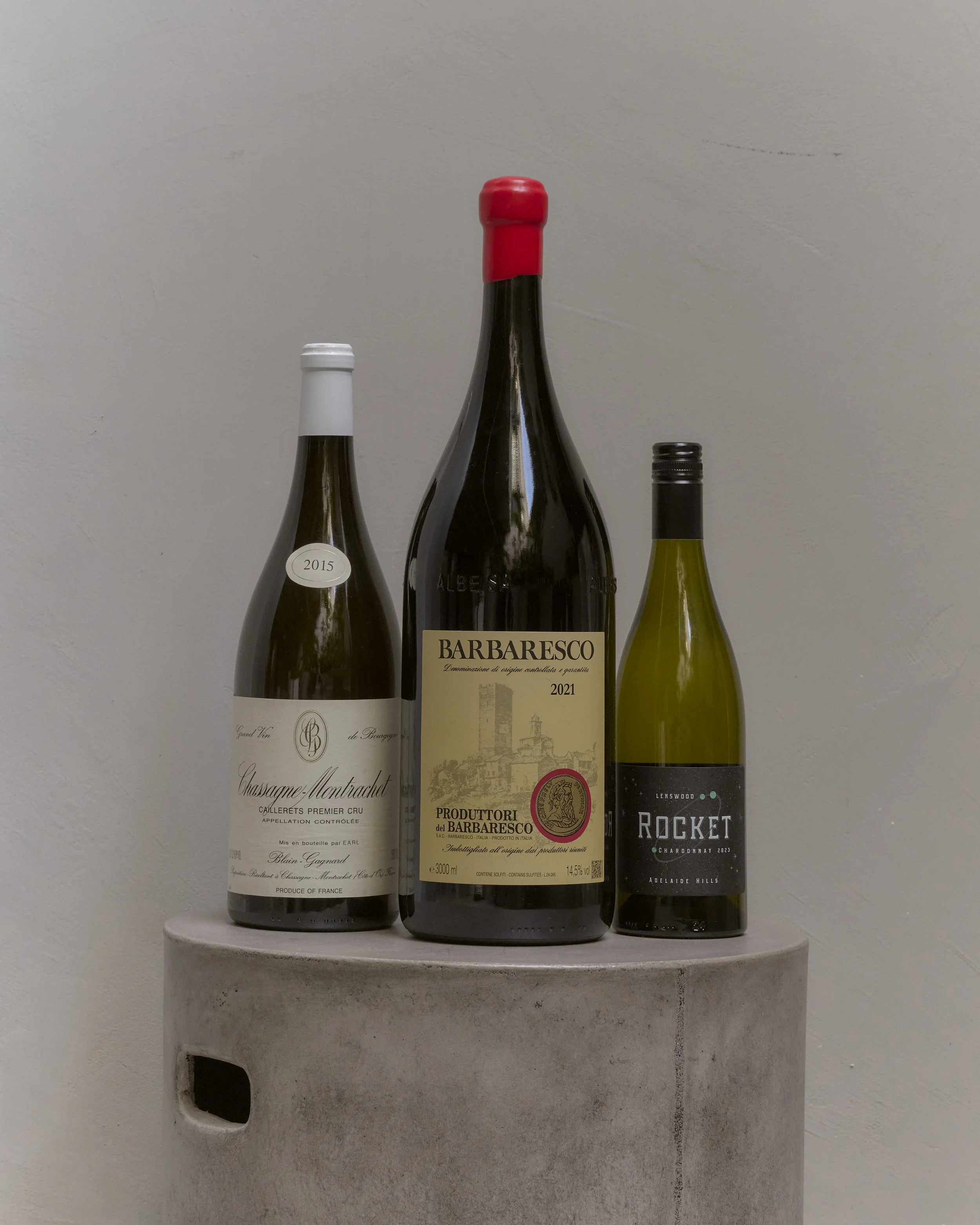 Three wine bottles are placed on a concrete pedestal against a plain wall background.