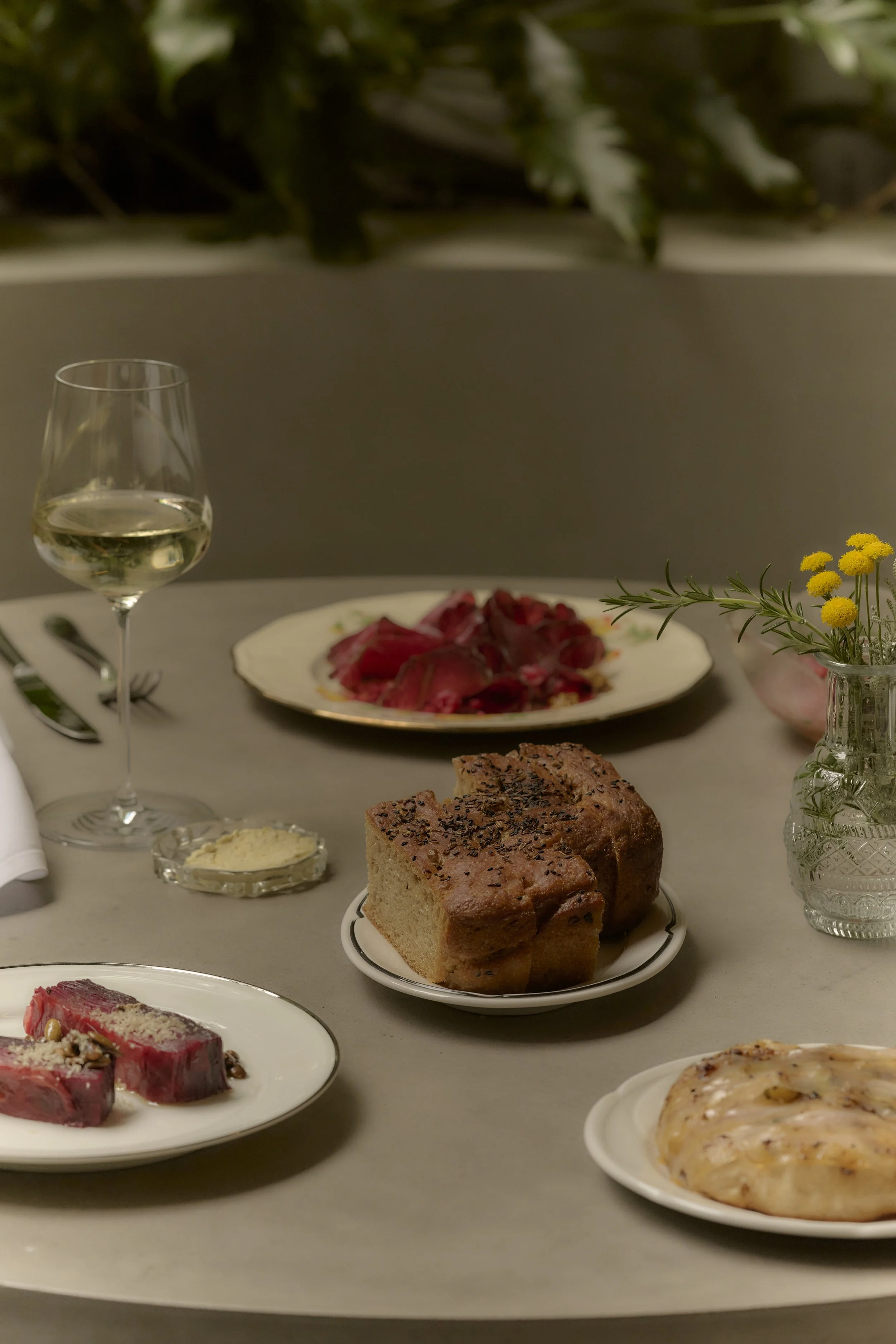 A dinner table set with a glass of white wine, slices of beef, loaf of bread, slices of cooked meat, and a flatbread, with a small jar of yellow flowers.