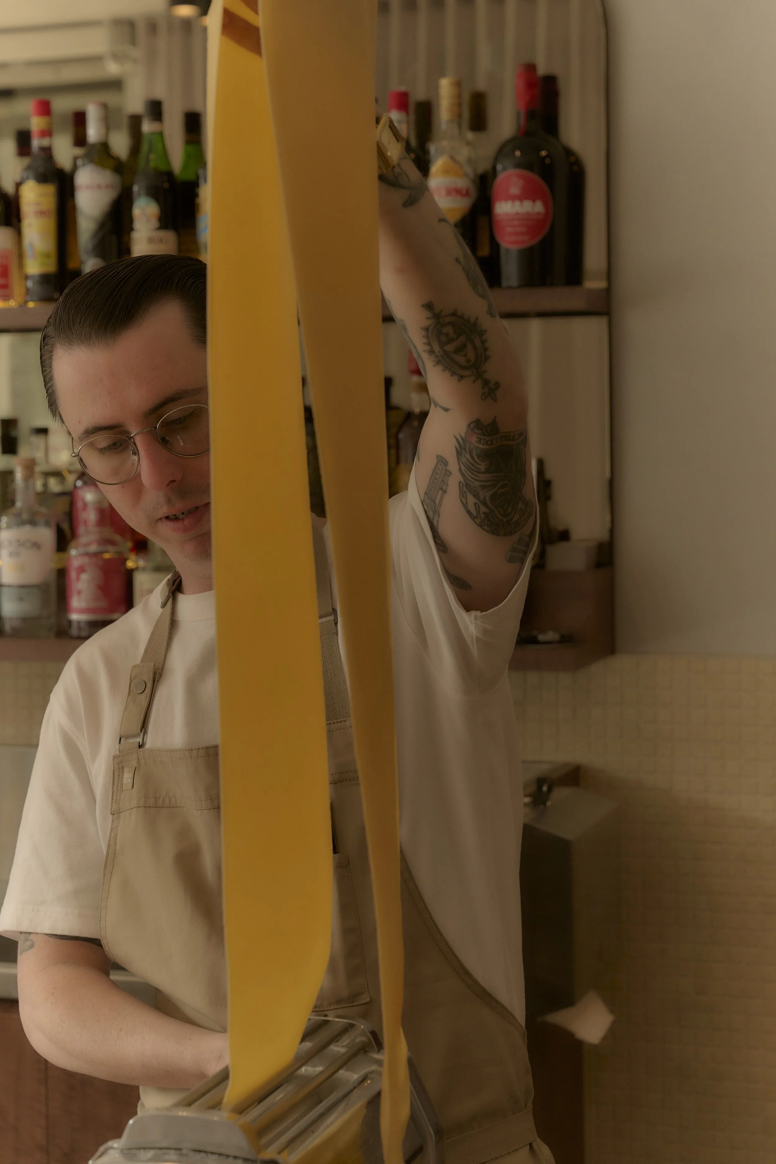 A person with glasses and tattoos on their arm cooking pasta in a kitchen. They are wearing a beige apron over a white shirt, and there are shelves with liquor bottles in the background.