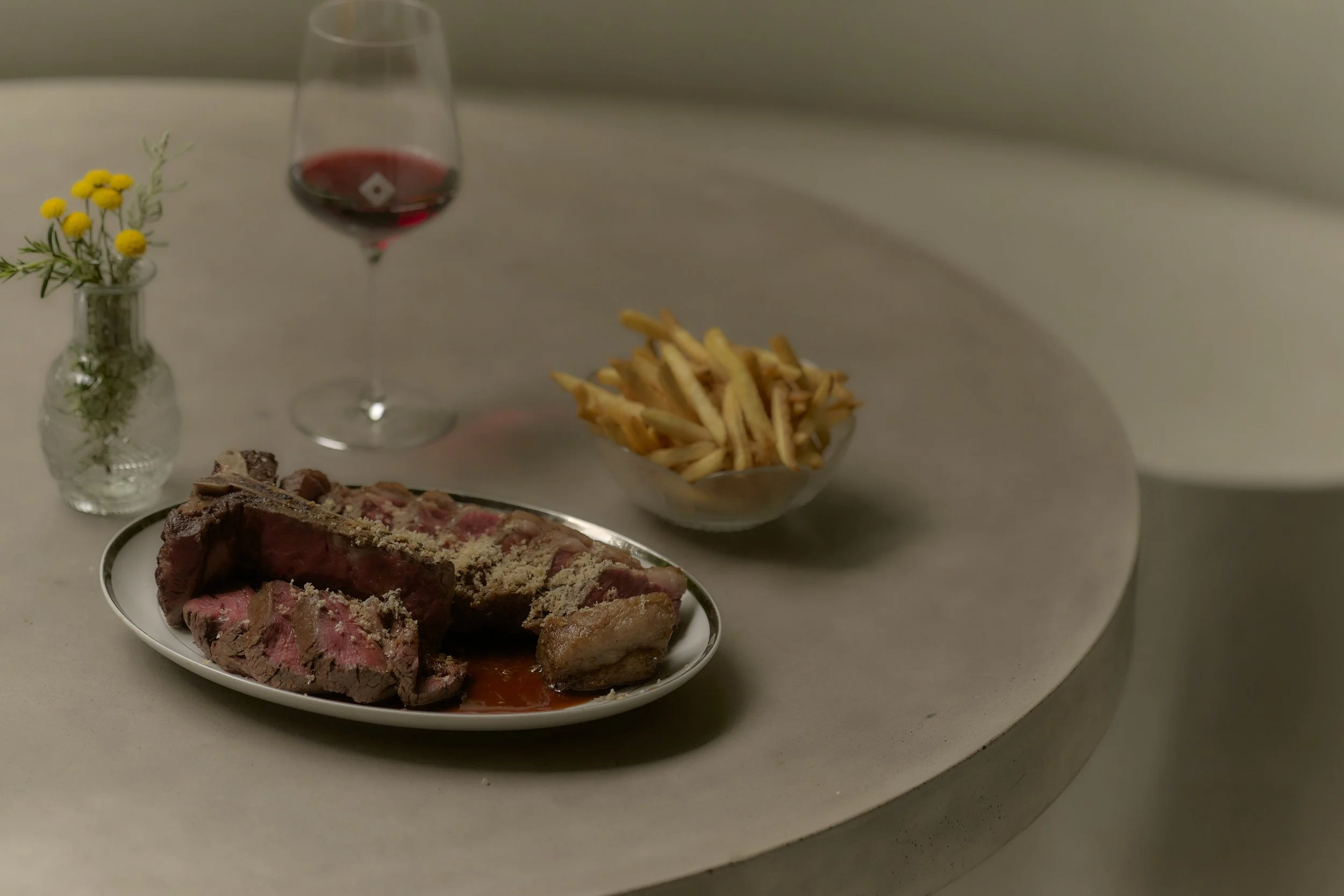 A meal with a plate of sliced beef steak, a bowl of French fries, a glass of red wine, and a small vase with yellow flowers on a marble table.