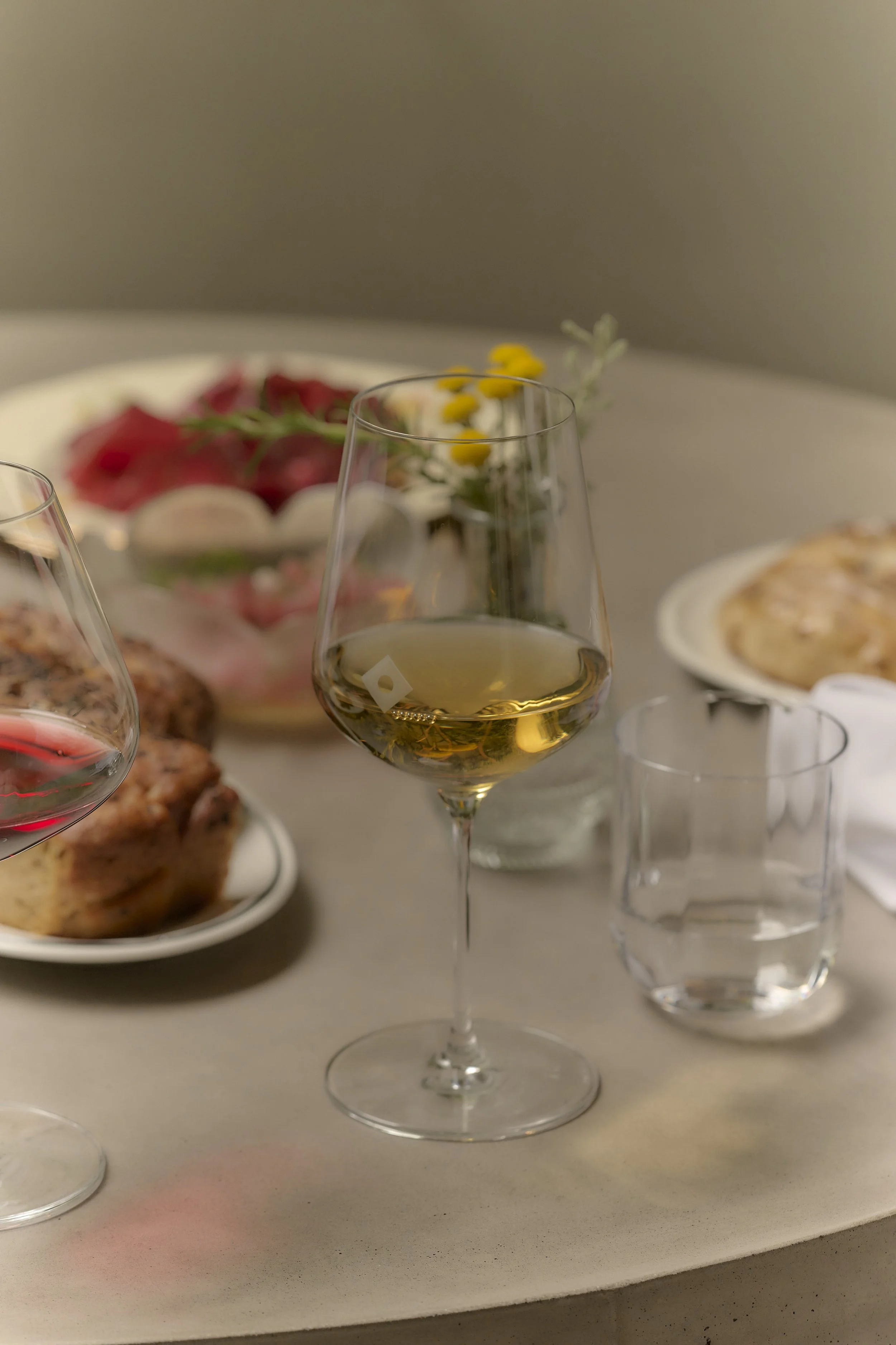 A dining table with a glass of white wine, a glass of red wine, a glass of water, a plate with a pie, a bowl of strawberries, a small vase with flowers, and some napkins.