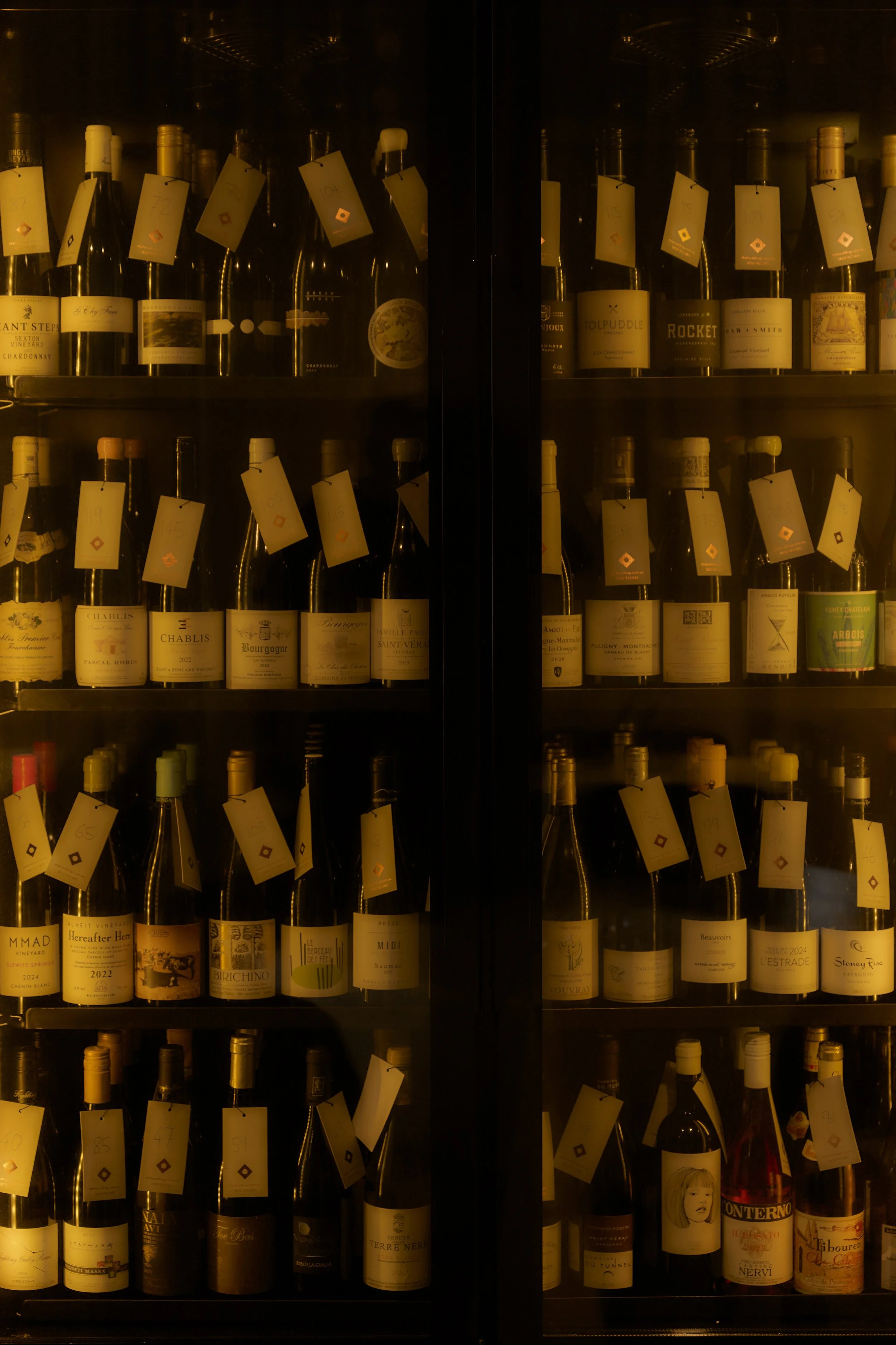 Glass wine cabinet filled with bottles of wine, each with a yellow tag attached, displayed on multiple shelves in a dimly lit room.