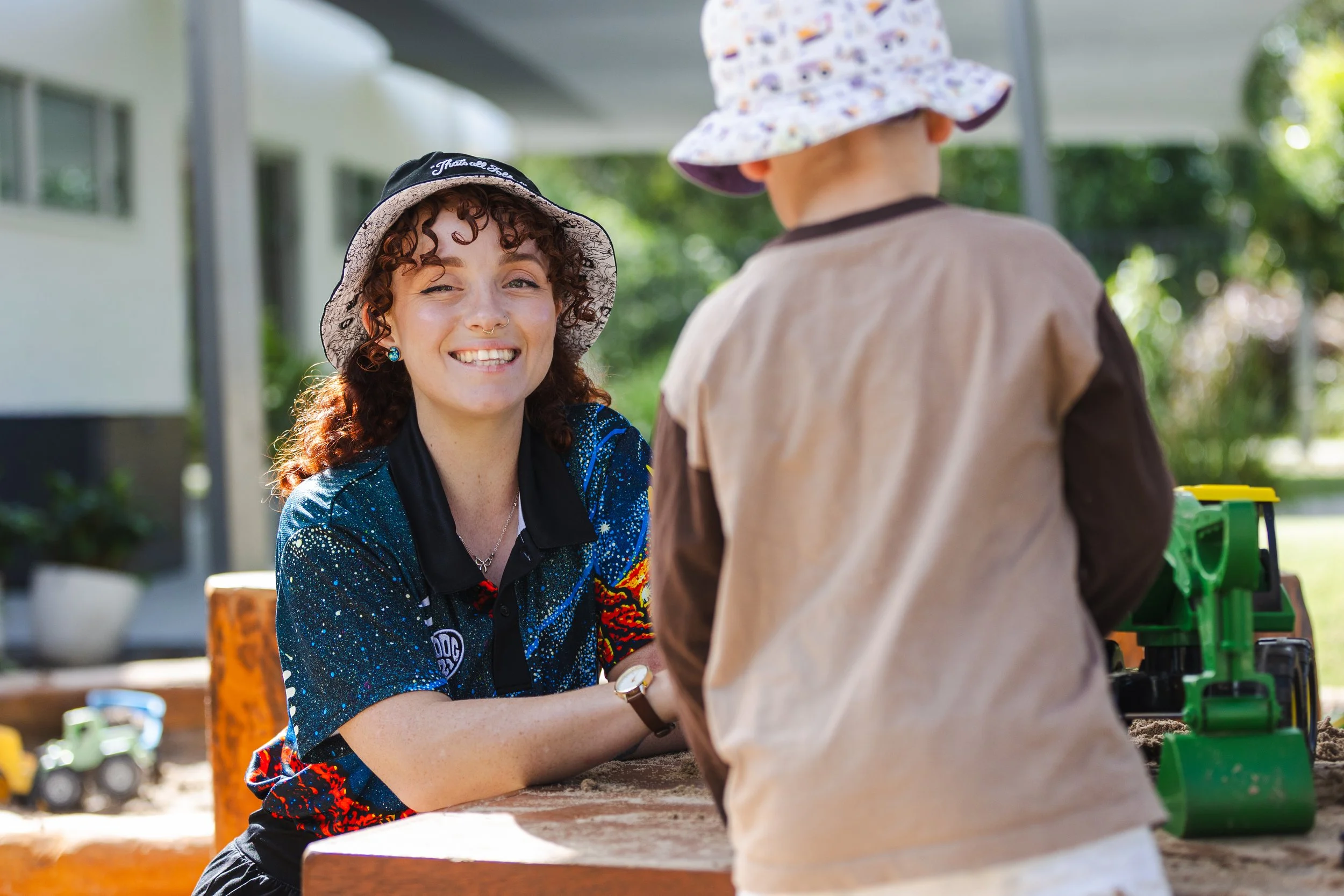 A woman with curly brown hair, wearing a camouflage bucket hat and a colorful, patterned sporty shirt, smiling and interacting with a child at an outdoor play area. The child is wearing a purple and white floral bucket hat and a beige jacket, facing away from the camera.
