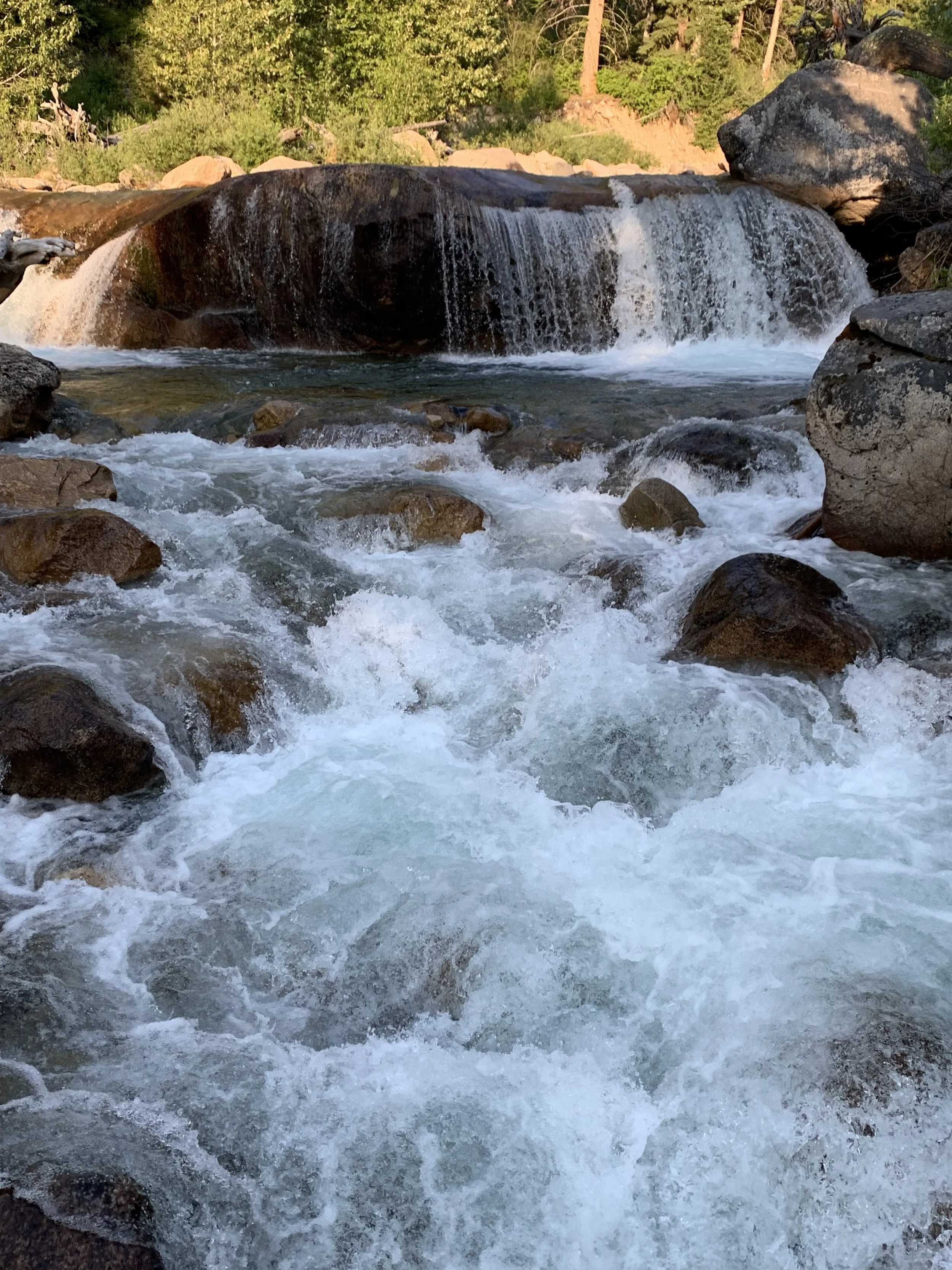 A river with rushing water flowing over rocks, with a small waterfall in the background, surrounded by trees and greenery.
