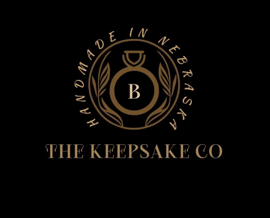A black background with a gold emblem and text. The emblem features a kettlebell with laurel branches and the letter B. Surrounding the emblem is the text 'Handmade in Nebraska'. Below, it says 'The Keepsake Co' in gold.