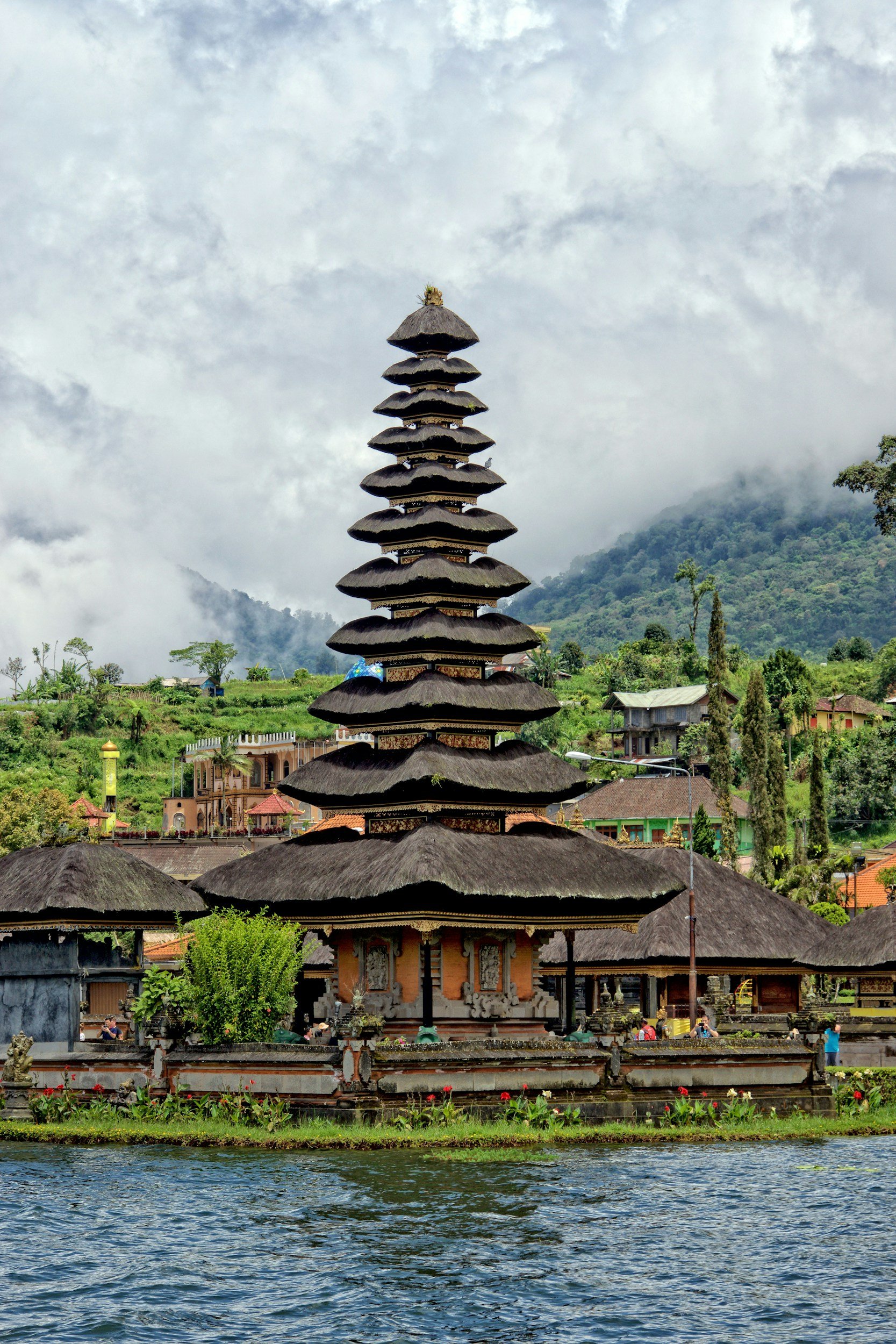 Bali temple with multiple layered thatched roofs, situated near a body of water with green landscape and mountains in the background, and cloudy sky overhead.