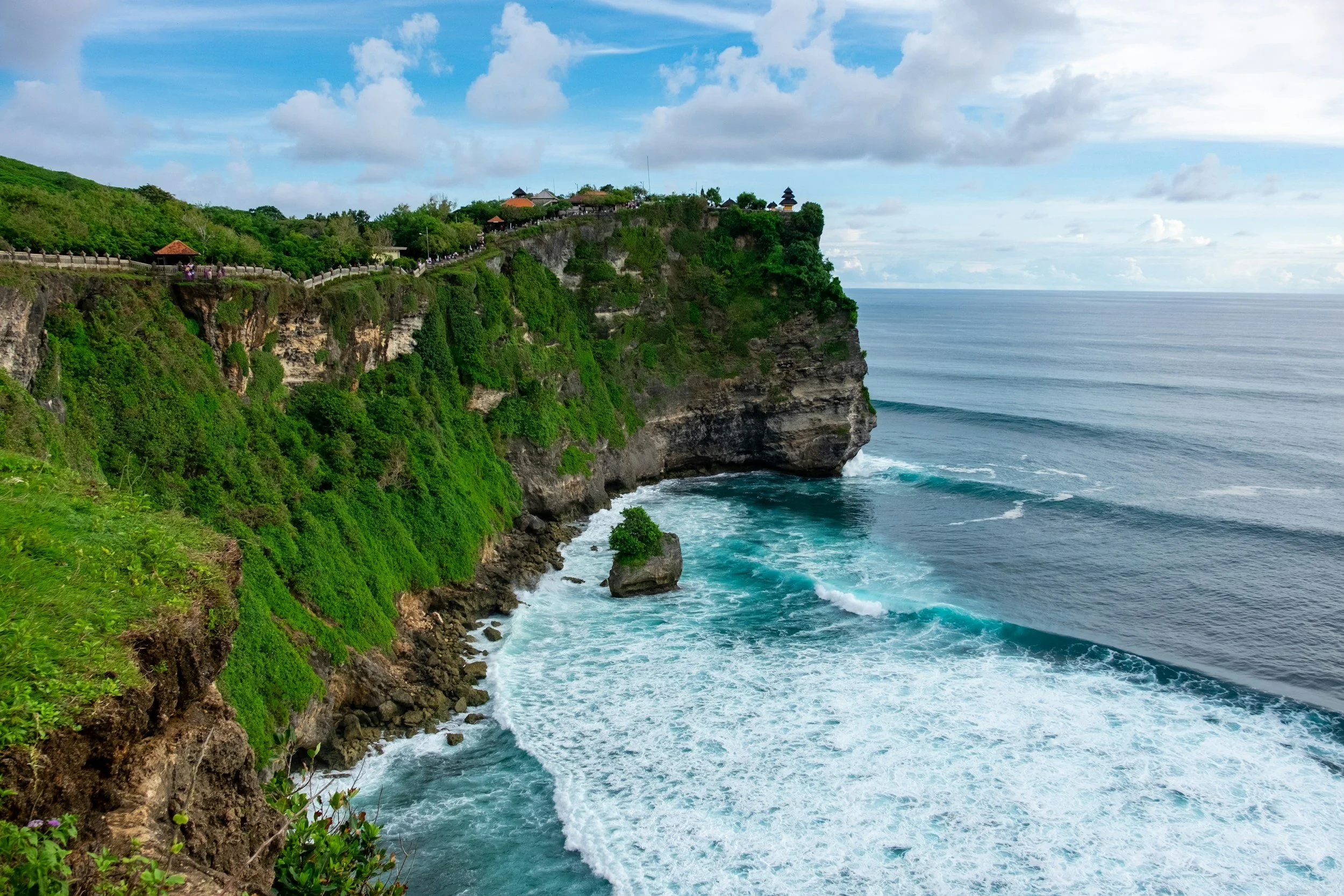 Uluwatu, Bali cliffside with lush green vegetation, rocky shoreline, and waves crashing against the coast, under a partly cloudy sky.