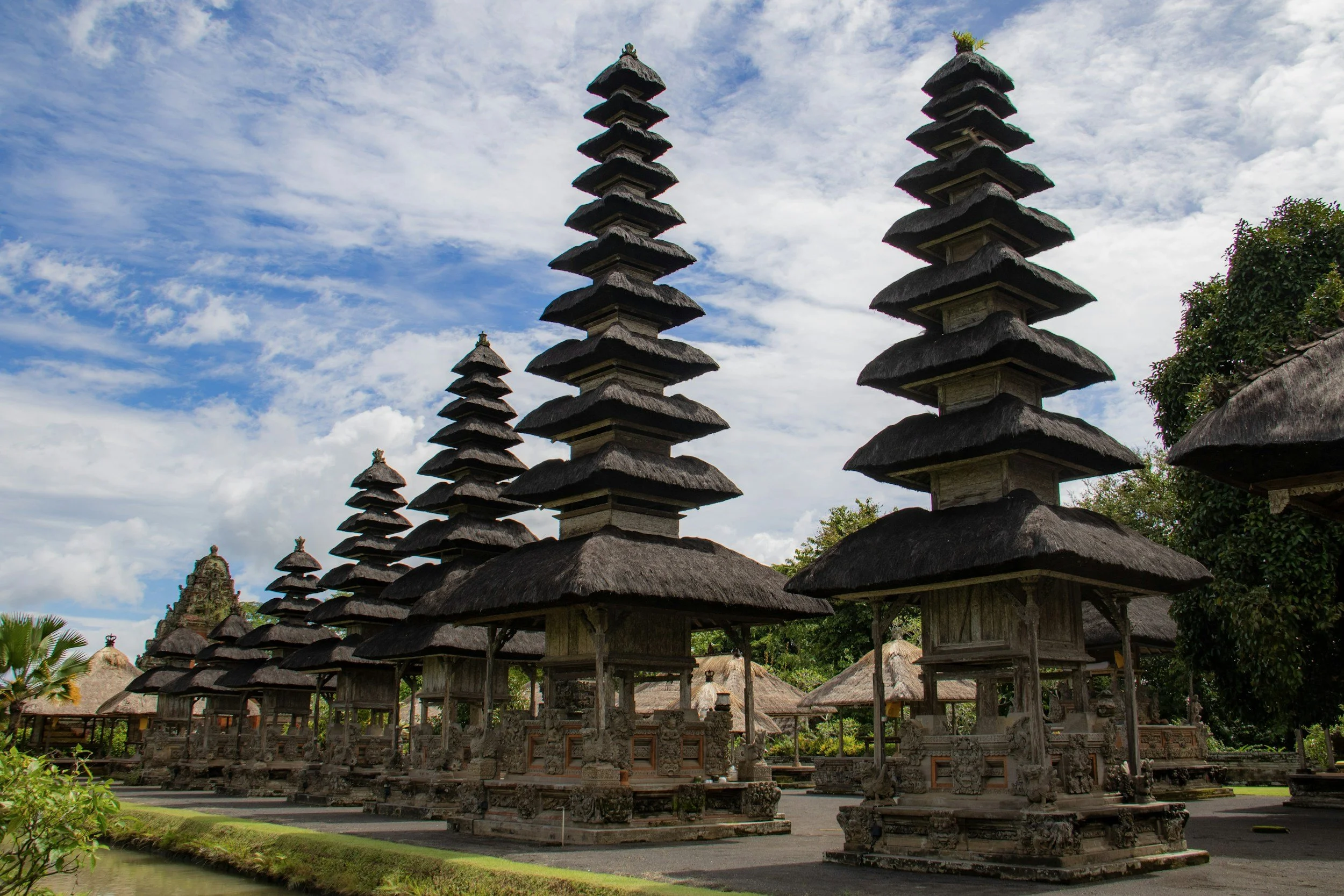 Traditional Balinese temple structures with multi-tiered thatched roofs in a lush green setting under a partly cloudy sky.