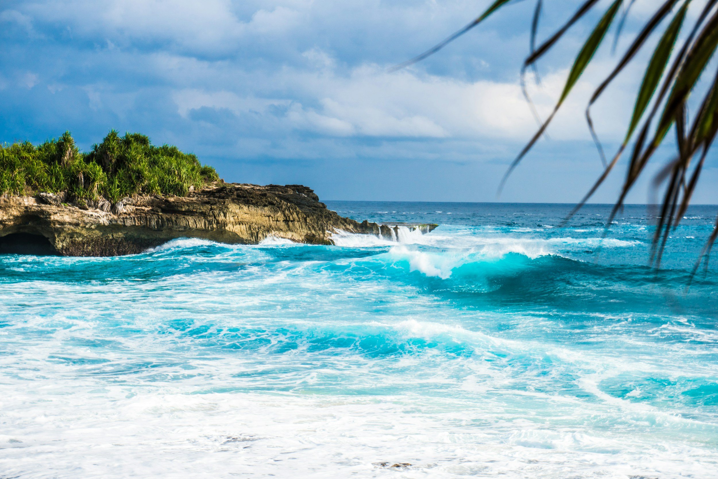 Ocean waves crashing against rocky shore with lush green vegetation and cloudy sky overhead.