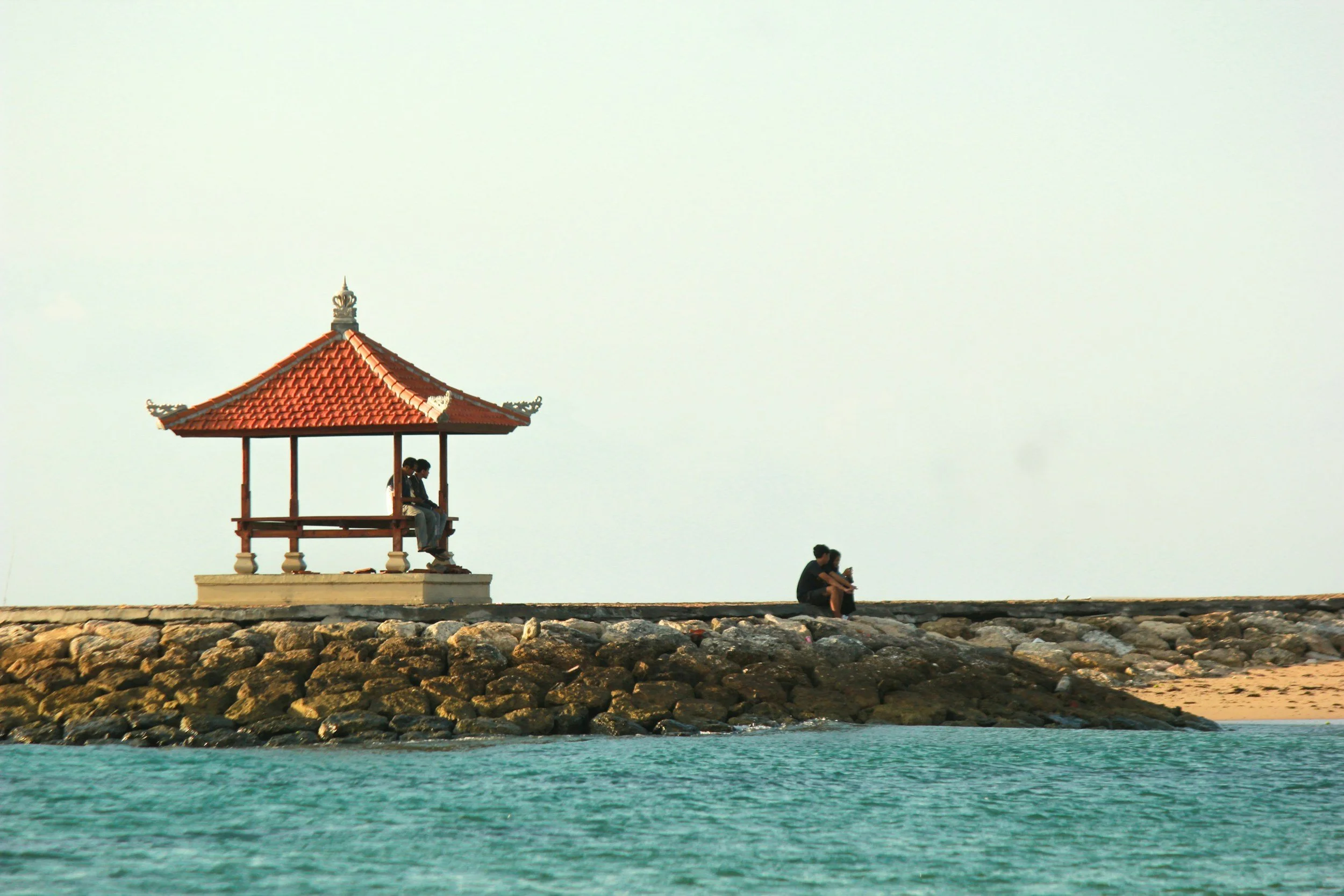 A rocky pier with a small pavilion with a red pagoda-style roof, two people sitting inside, and two people sitting on the rocks nearby, with turquoise water in the foreground and an open sky in the background. Located in Sanur, Bali, Indonesia.