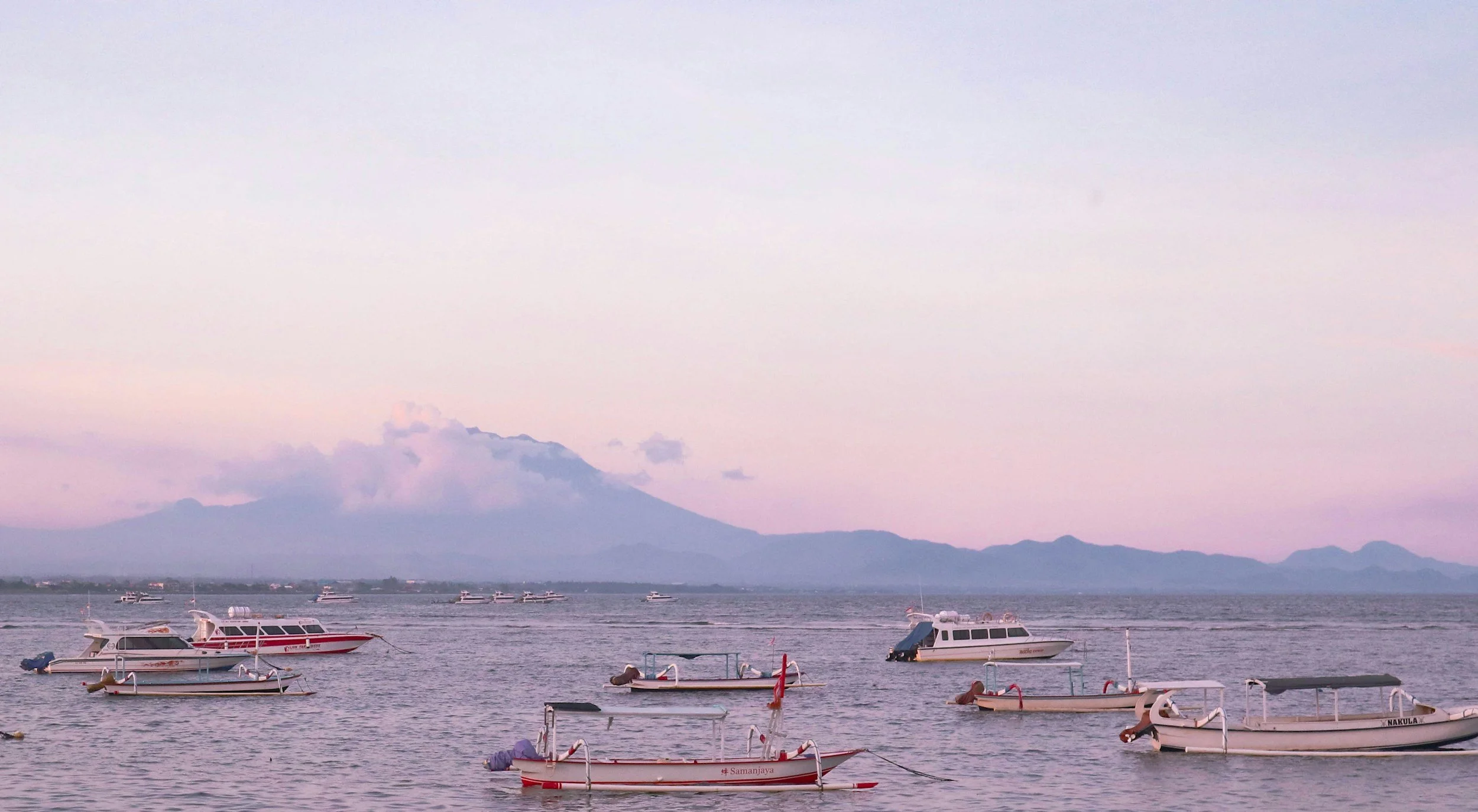 Boats floating on a calm body of water with a mountain and a pinkish-purple sky in the background.