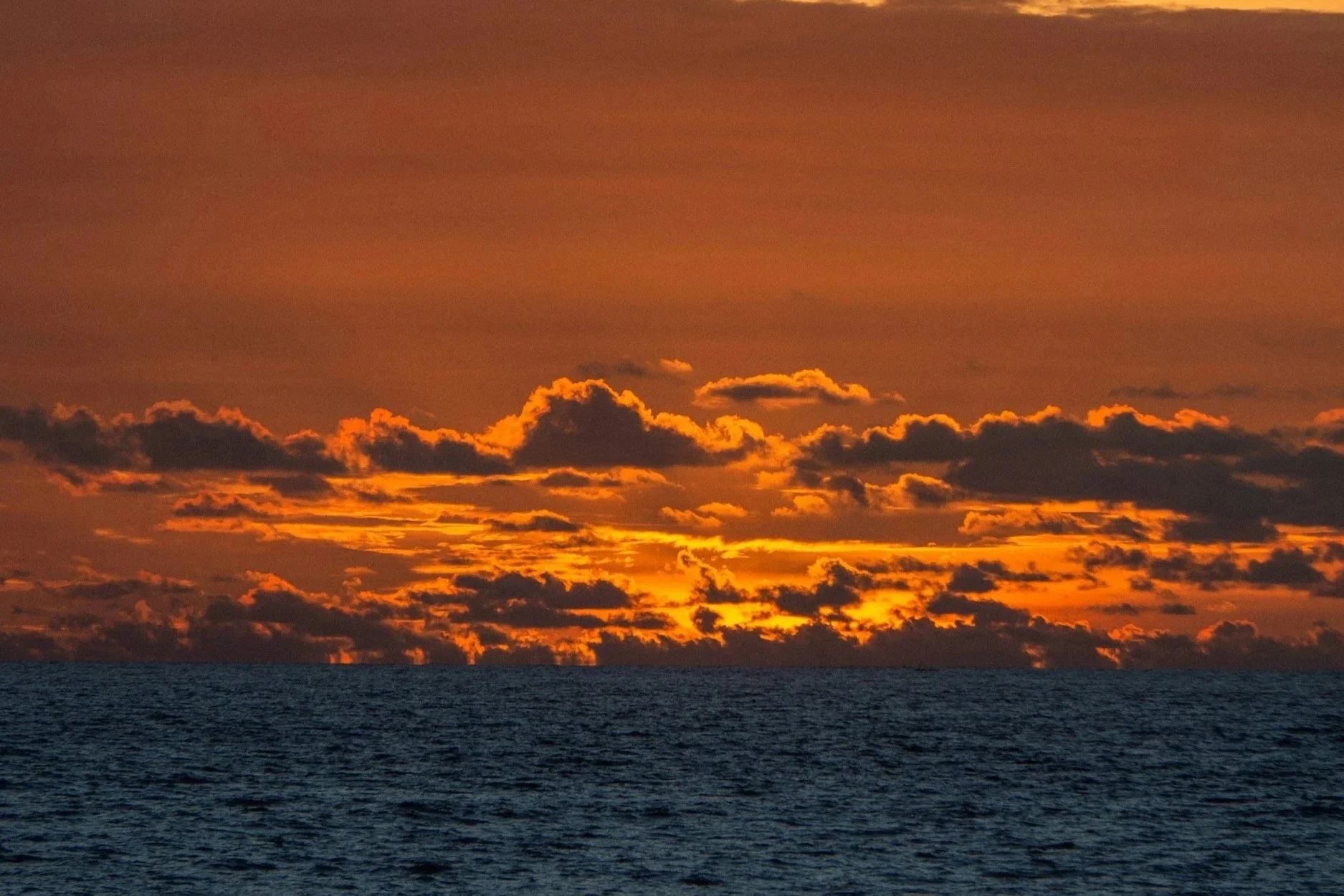 Sunset over the ocean with a partly cloudy sky emitting orange and yellow hues, calm water in the foreground.