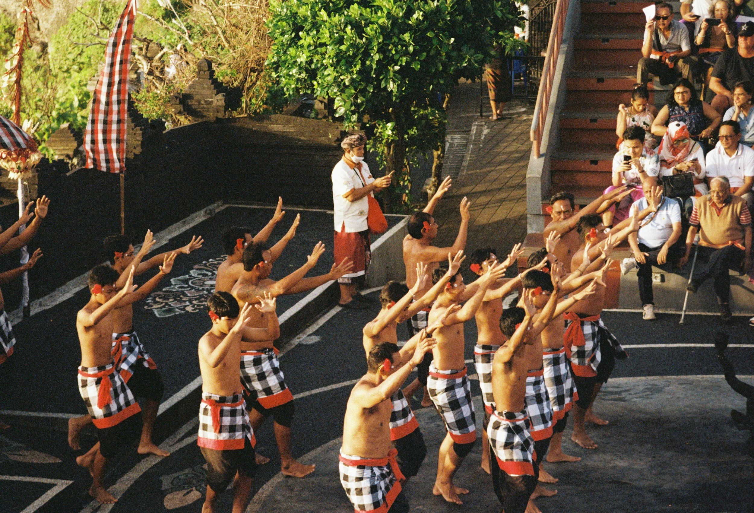 Group of men performing a traditional Balinese dance, wearing checkered sarongs and headbands, during a cultural event with spectators seated on steps and standing nearby.
