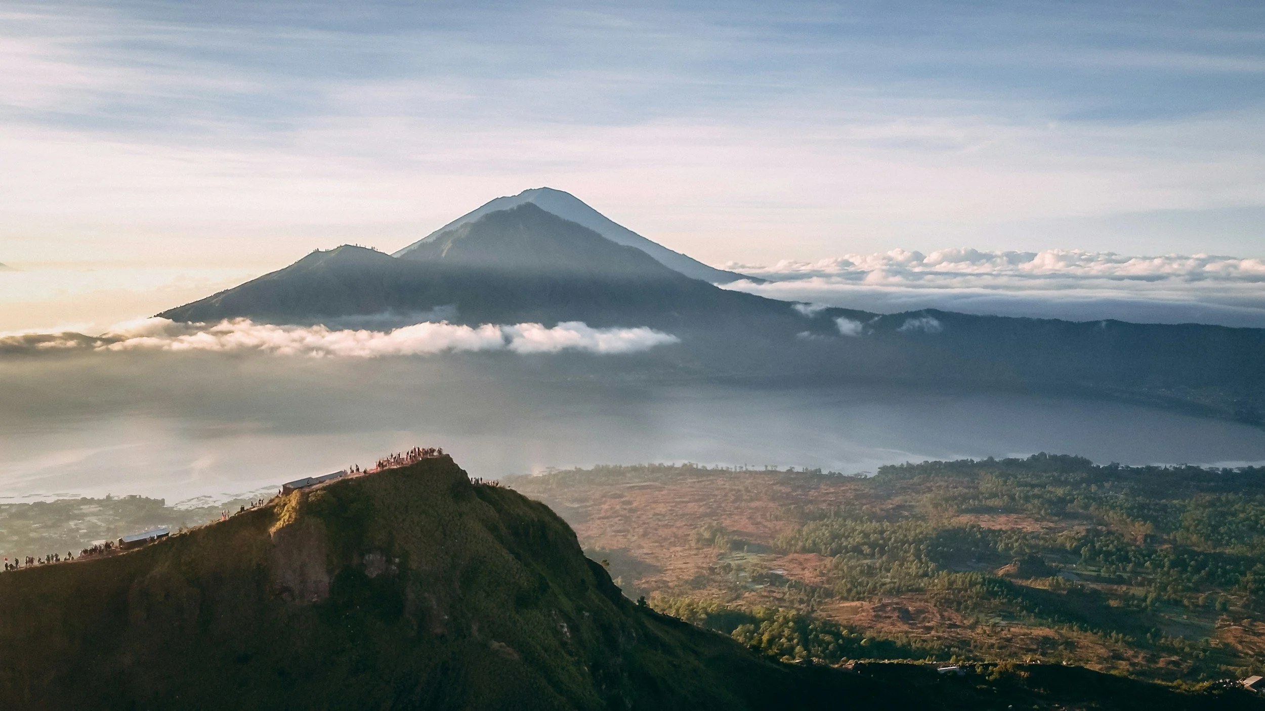Scenic view of a large mountain with a volcano, partially covered by clouds, behind a smaller green hill with a group of people watching from the top, during daytime with partly cloudy sky.