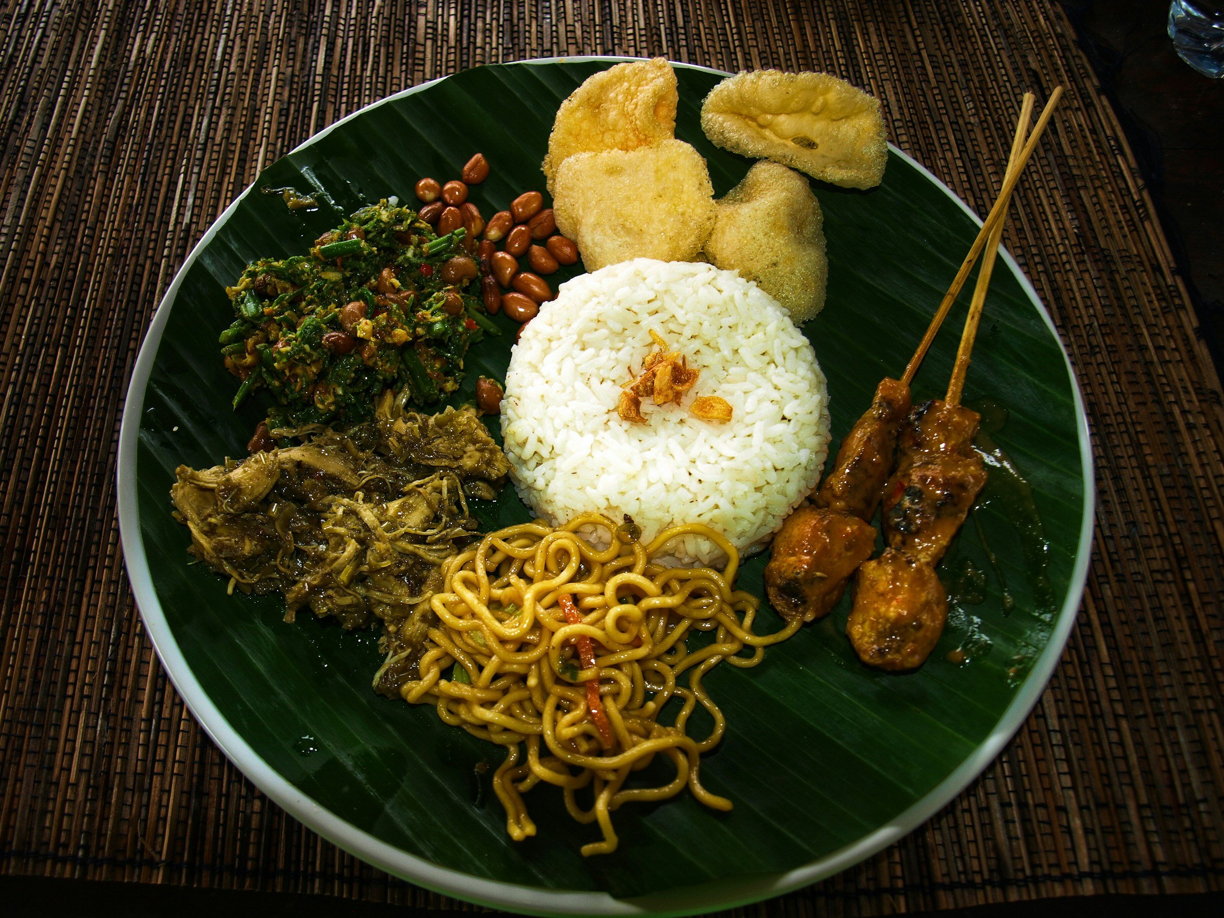 Plate of Indonesian cuisine with rice, noodles, fried vegetables, and skewered meat, served on a green banana leaf.