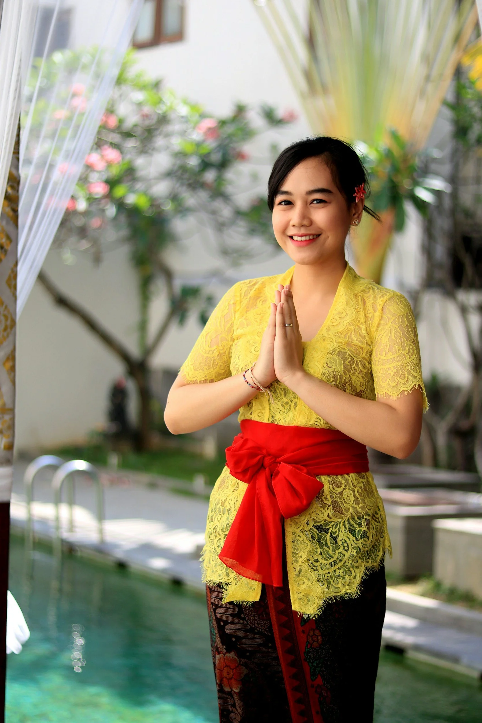 A young Balinese woman dressed in traditional Indonesian attire standing outdoors beside a pool, smiling and holding her hands together in a respectful gesture.