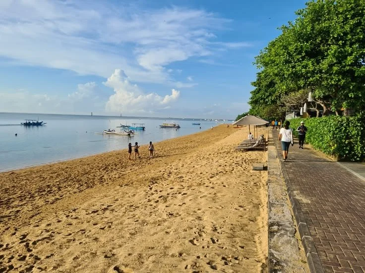 Sanur beach with boats in the water, people walking on the sandy shore, and a walkway lined with trees and benches.