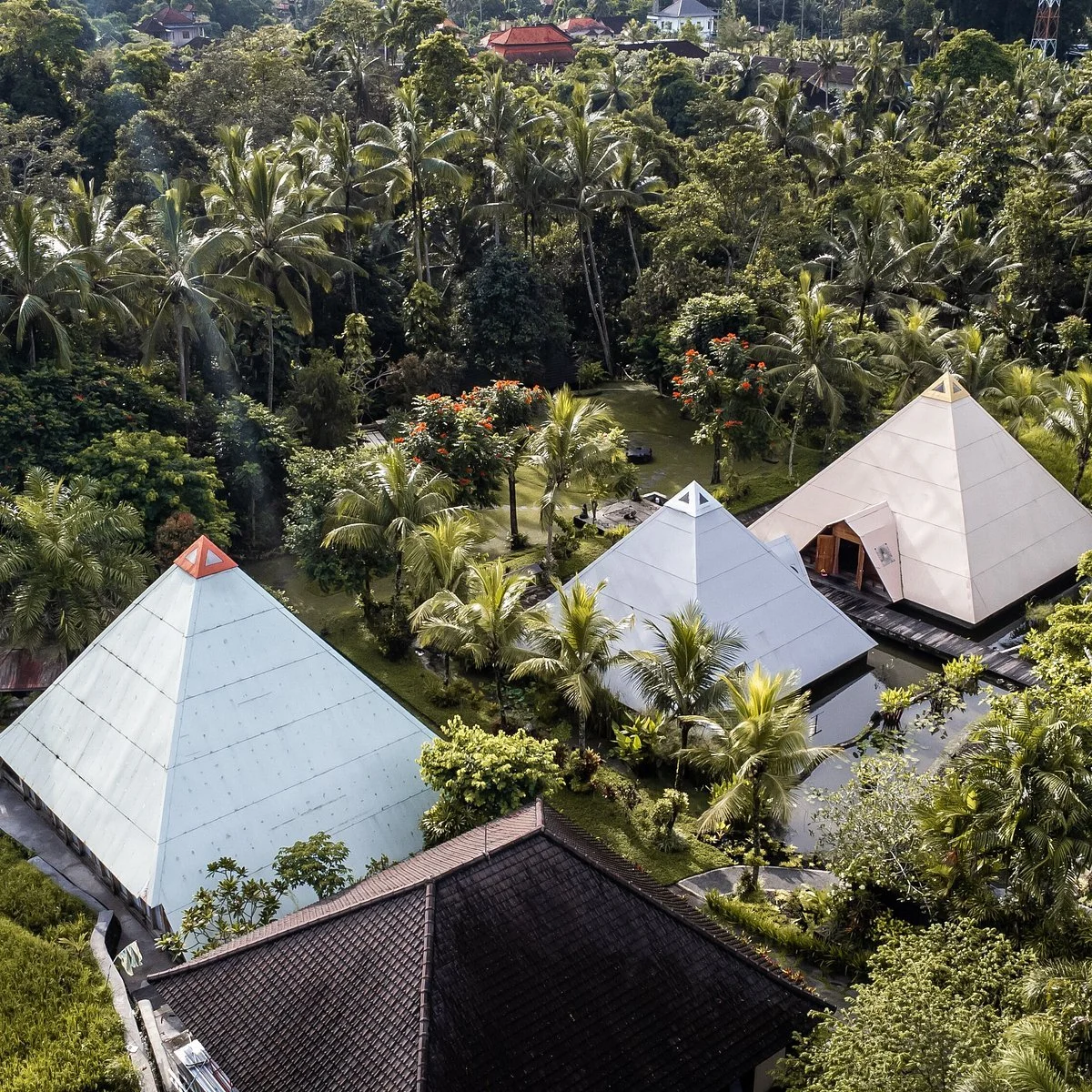 An aerial view of a tropical landscape with three white pyramid-shaped buildings surrounded by lush green trees and plants located in Bali at Pyramids of Chi.