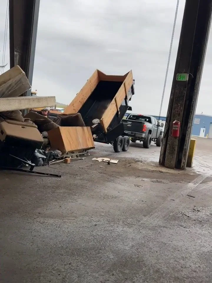 A partially tipped black dump trailer with a wooden frame, loaded with drywall, inside an industrial warehouse with an open garage door, a truck, and various construction debris scattered on the concrete floor.