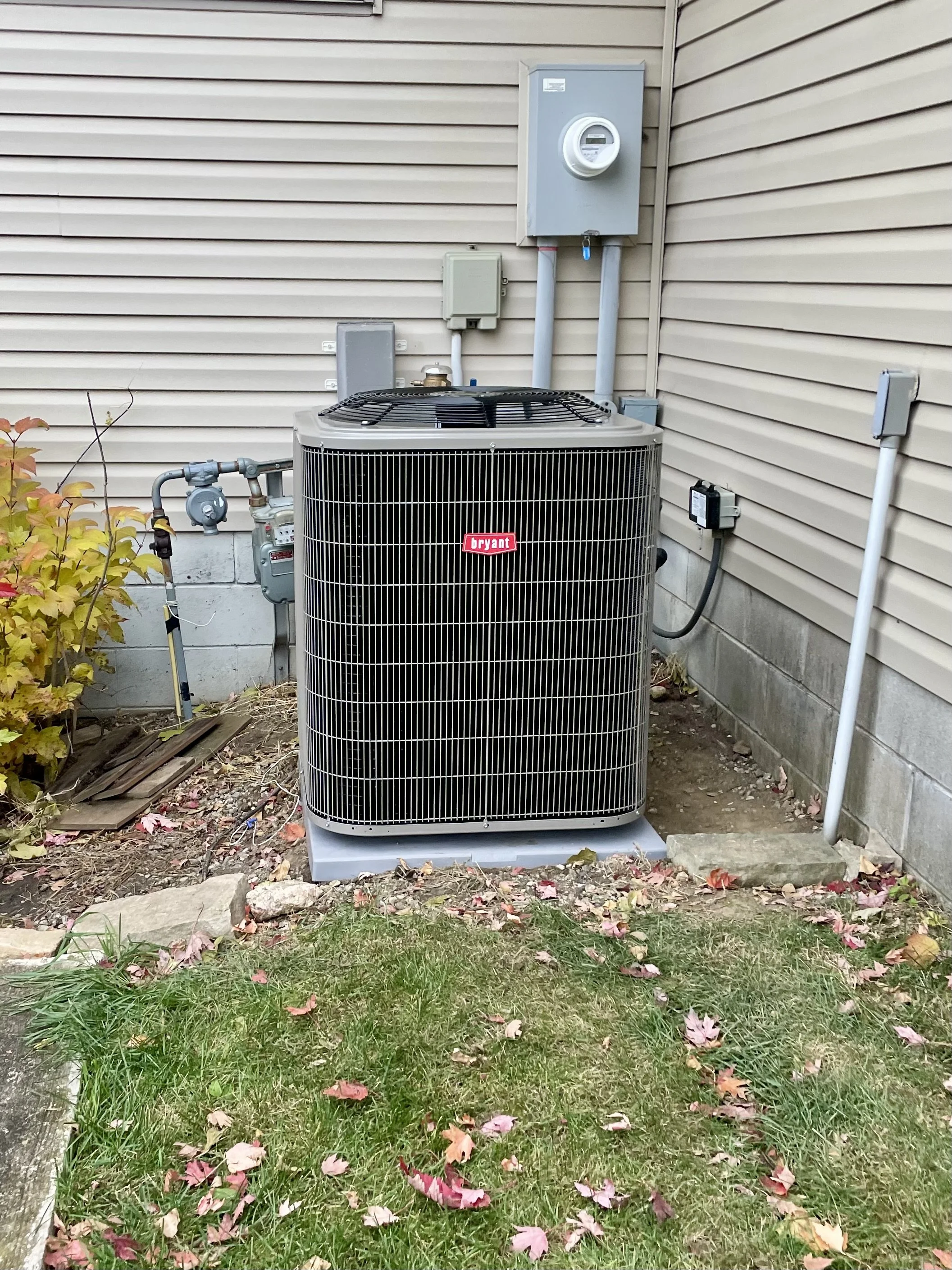 An outdoor air conditioning unit next to a house with beige siding, surrounded by grass, leaves, and small landscaping stones.