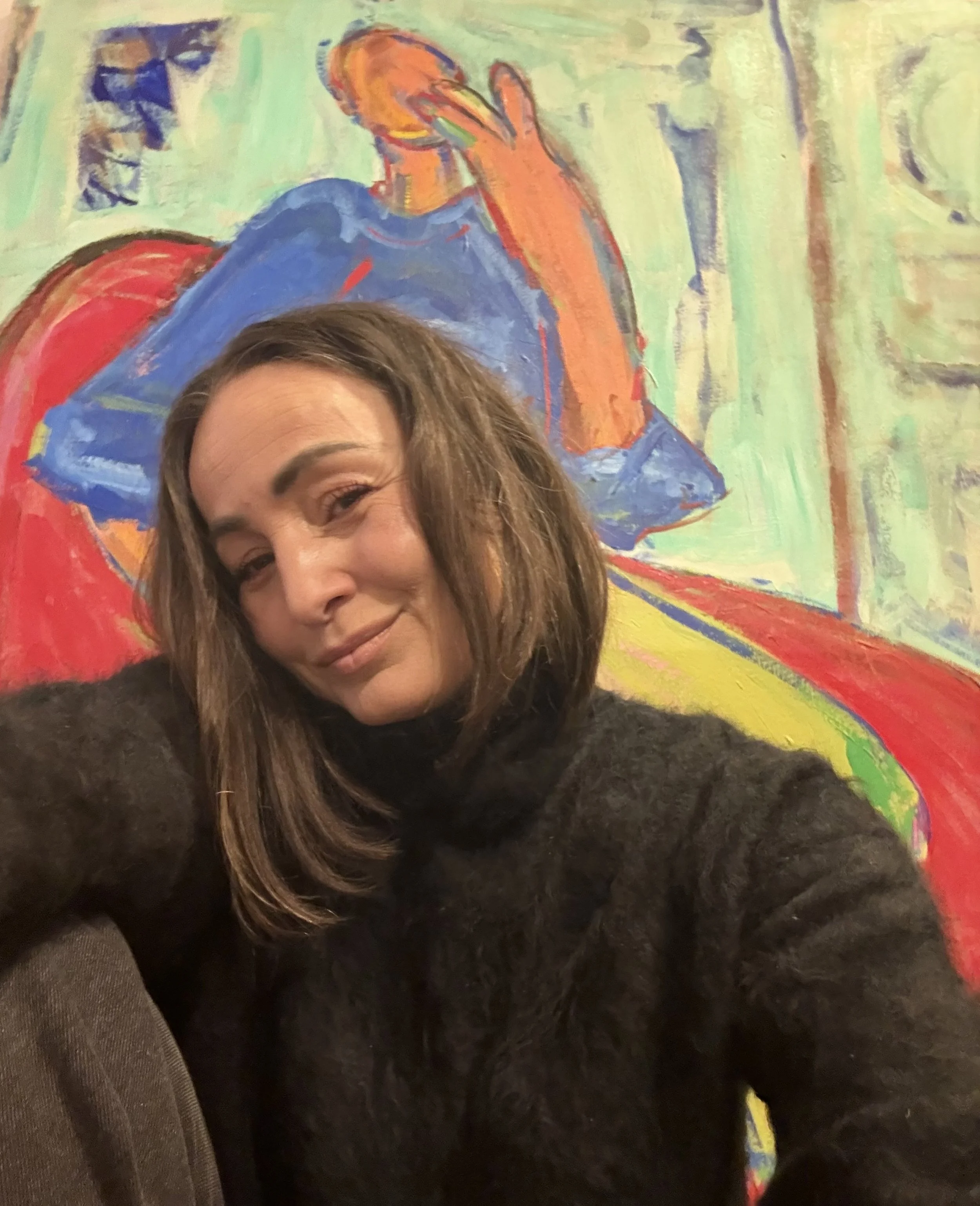 A woman with shoulder-length brown hair smiling at the camera, sitting in front of a colorful, abstract painting of a person with orange hair, a blue dress, and a red and yellow background.