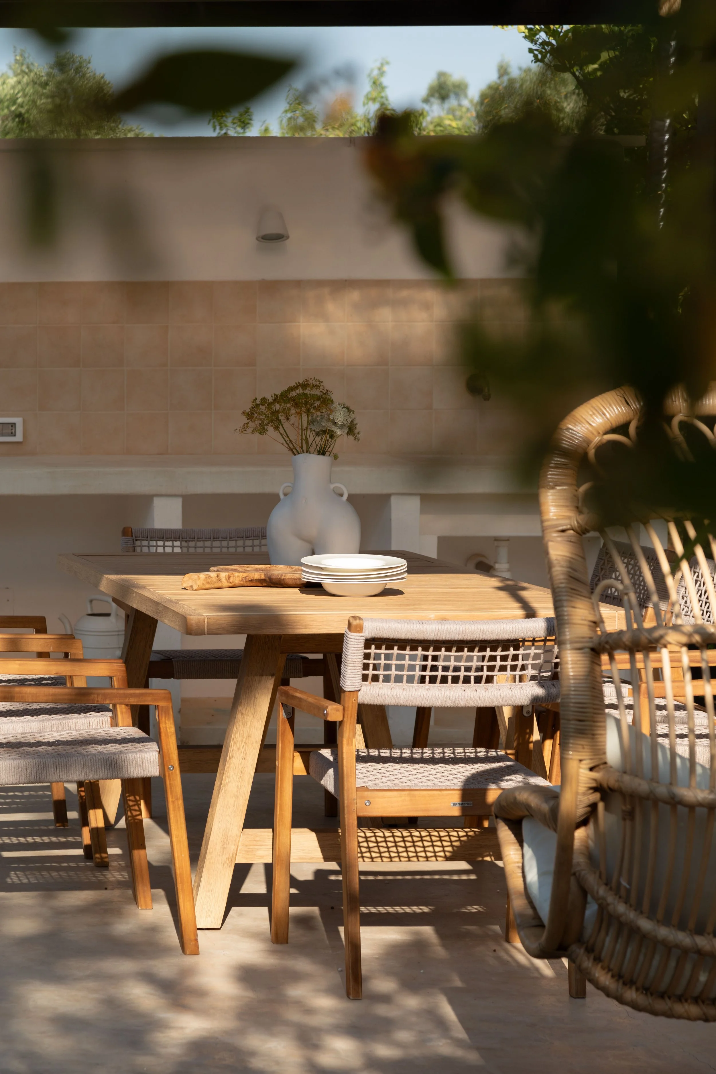 Outdoor dining area with a wooden table, surrounded by rattan and wooden chairs, a large white vase with flowers in the background, and sunlight creating shadows on the floor.