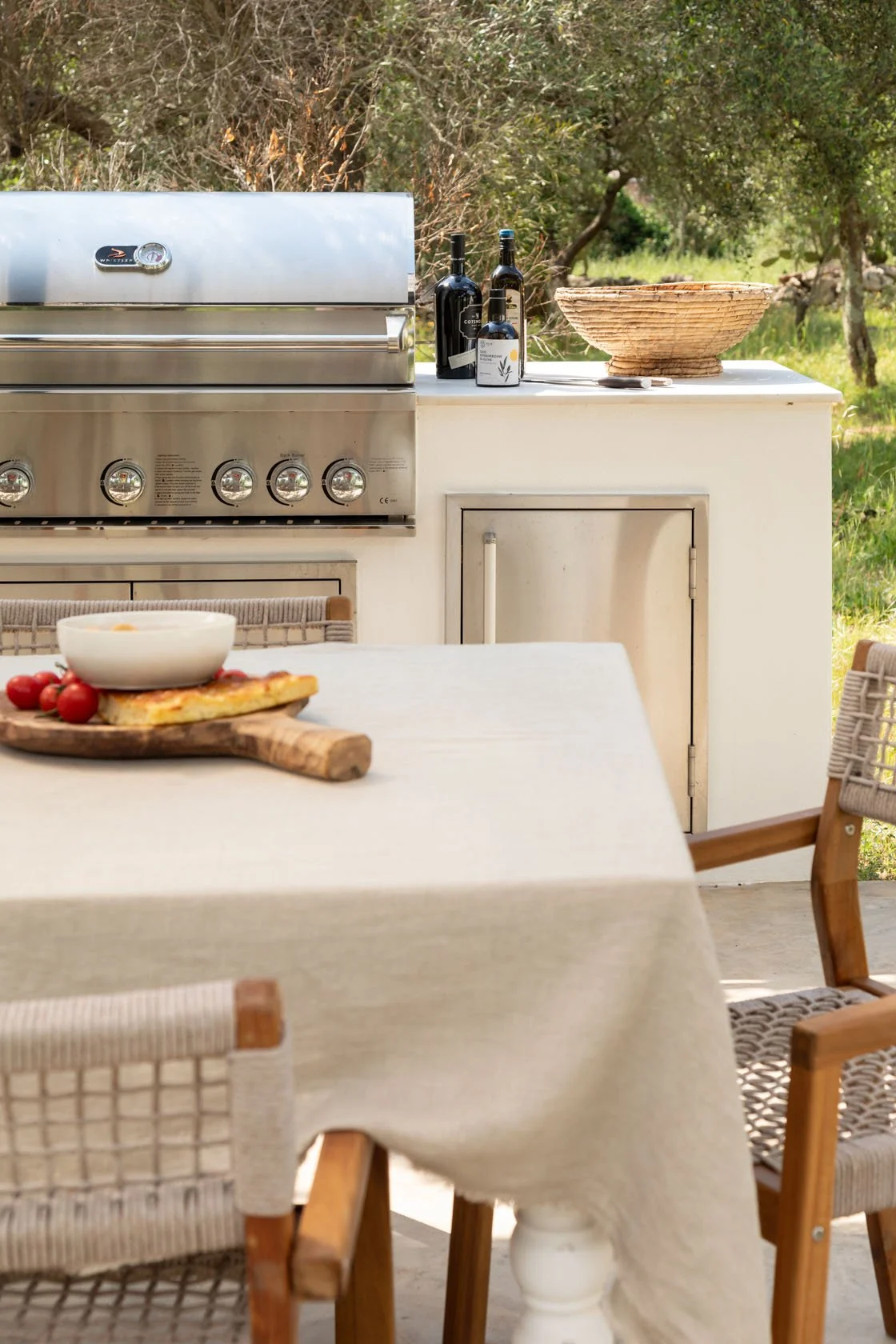 An outdoor kitchen with a grill, bottles of wine, a woven basket, a table with a pizza and cherry tomatoes, and chairs, set in a green, wooded area.